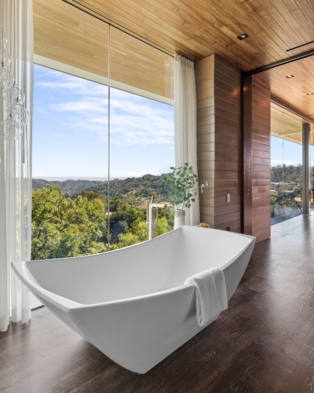 The modern primary bathroom at our Benedict Canyon House takes in views of L.A.'s rolling hillsides through floor to ceiling glass walls. Photo by @mrbarcelo