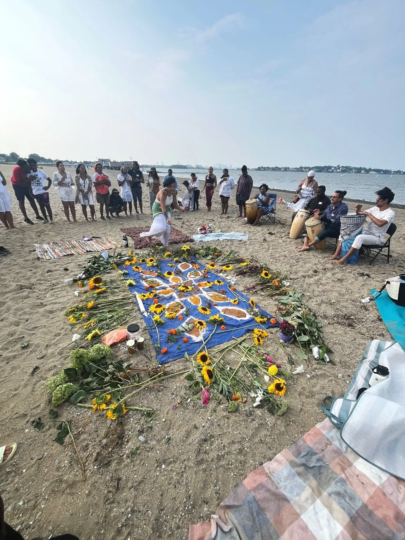 This is a picture of people gathered around sunflowers, art, and dyed linen on the beach.