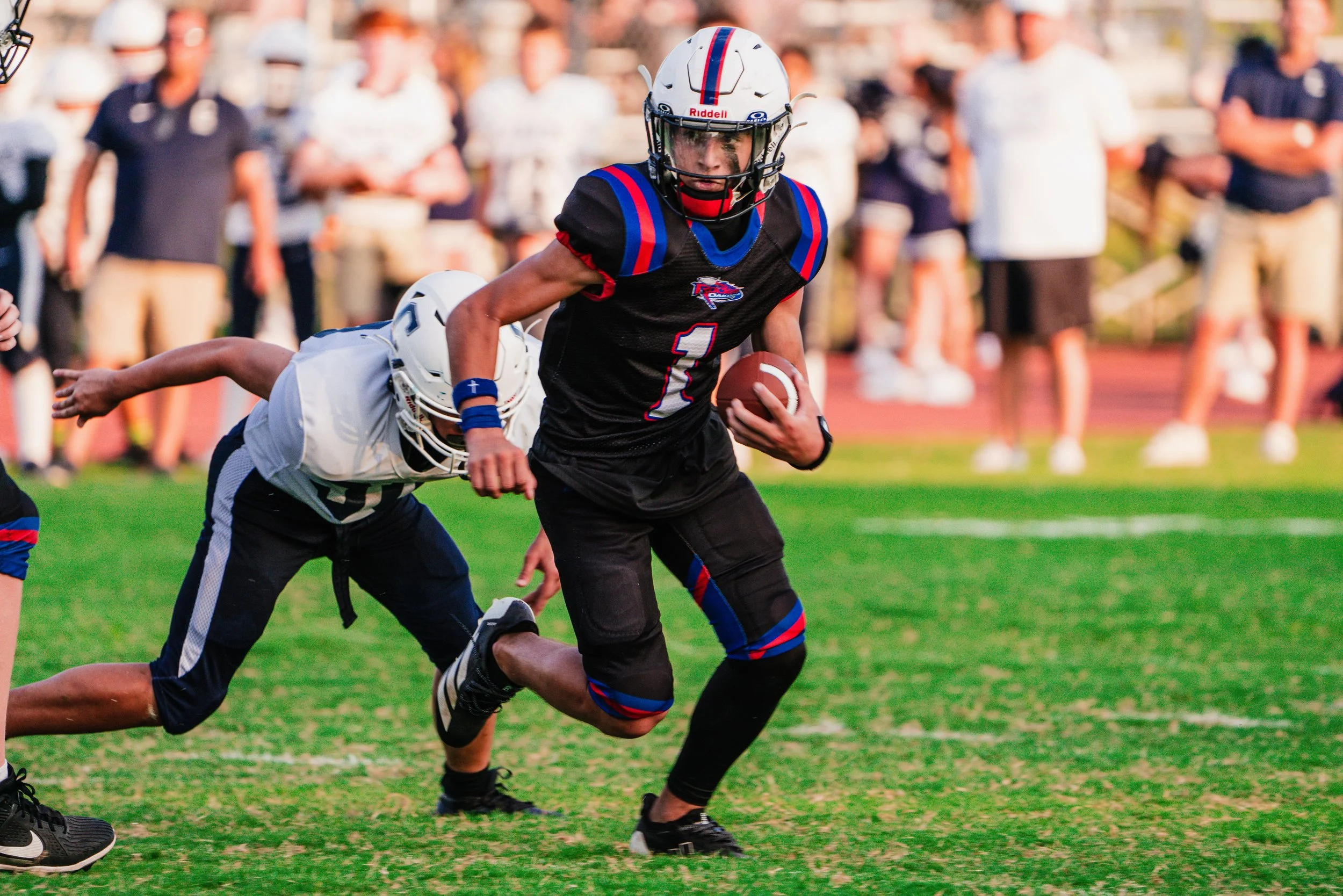 A boy in a football uniform running with a football while a player falls behind him on the field, with spectators blurred in the background.