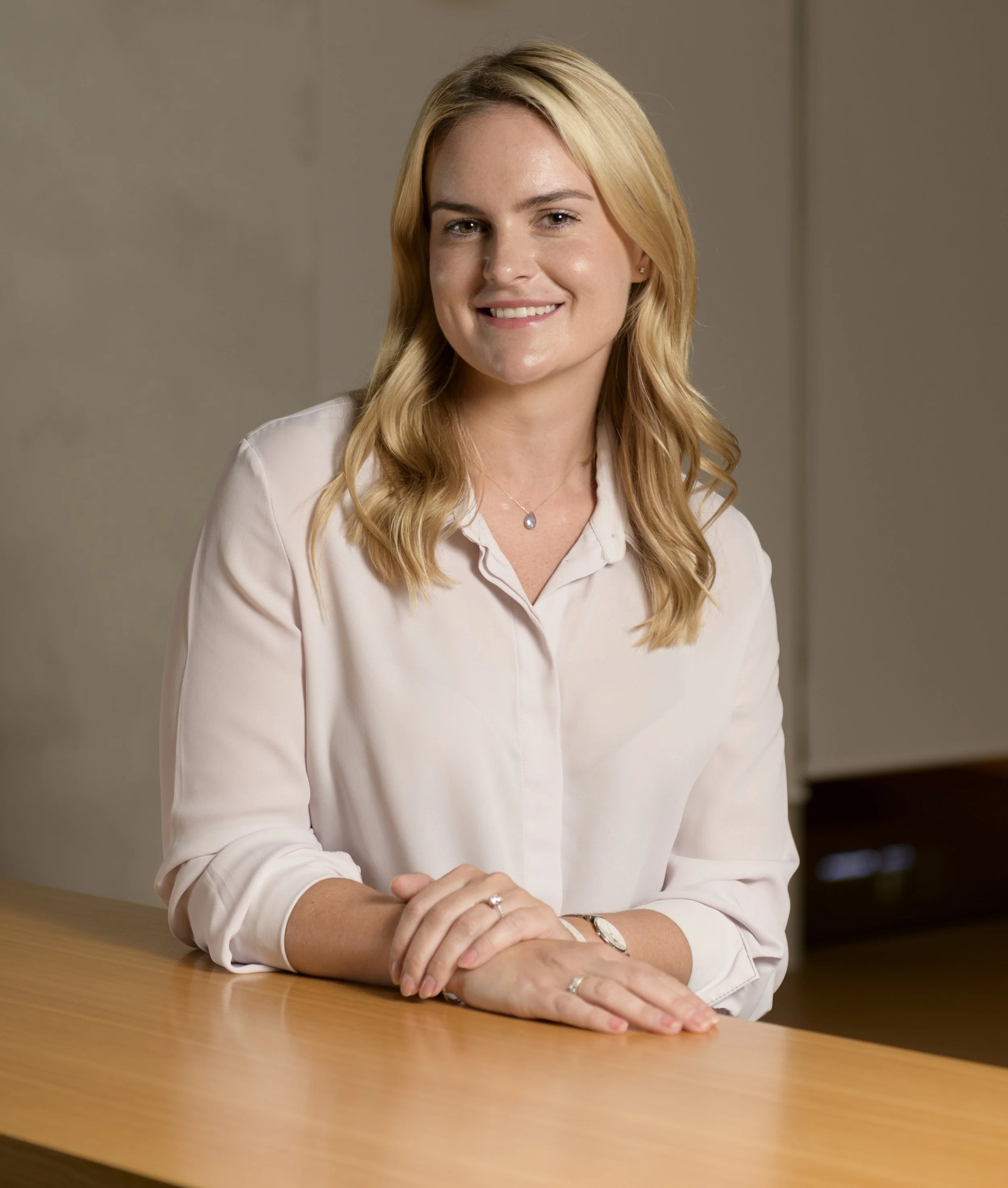 Woman with blonde hair wearing a white blouse, sitting at a wooden table, smiling.