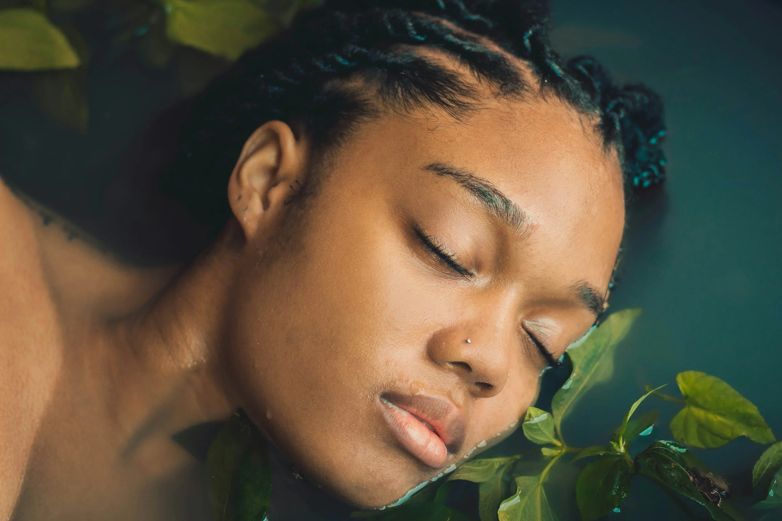 A woman with dark skin and braided hair is lying in water surrounded by green leaves, with her eyes closed and a peaceful expression.