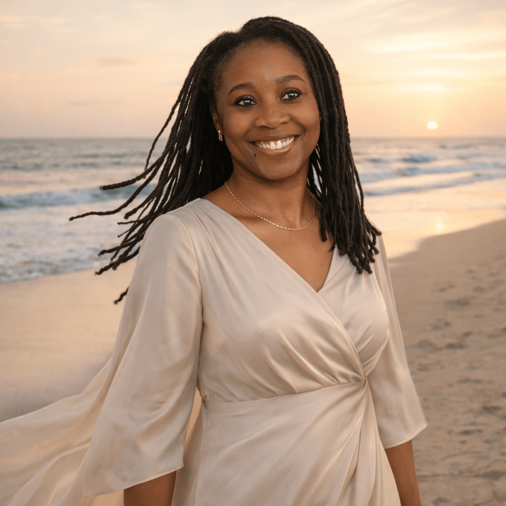 A woman with dreadlocks smiling on a beach at sunset, wearing a light-colored dress.