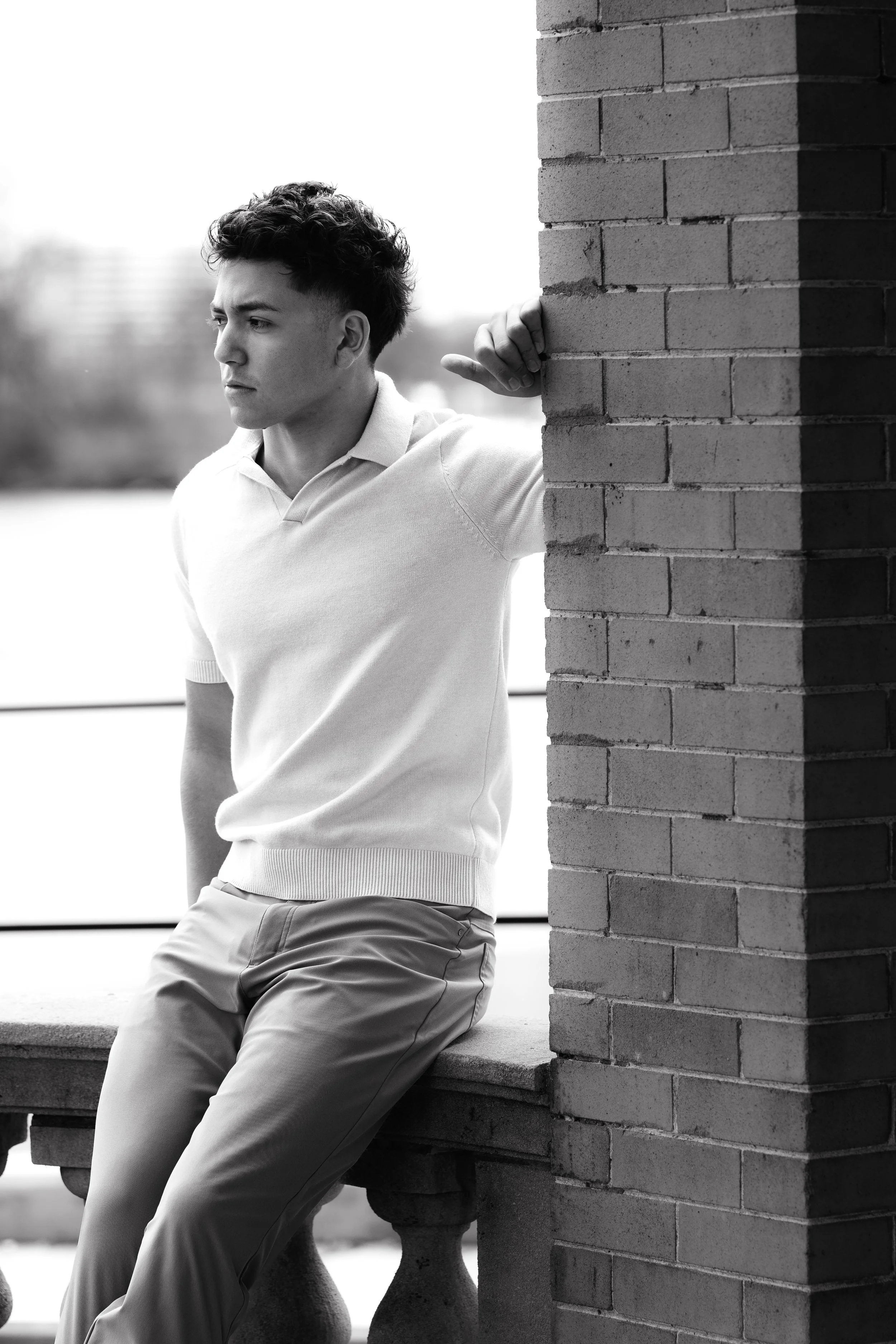 A young man with curly hair, wearing a light-colored polo shirt and pants, leaning against a brick wall on a bench outdoors, looking pensively into the distance in black and white.