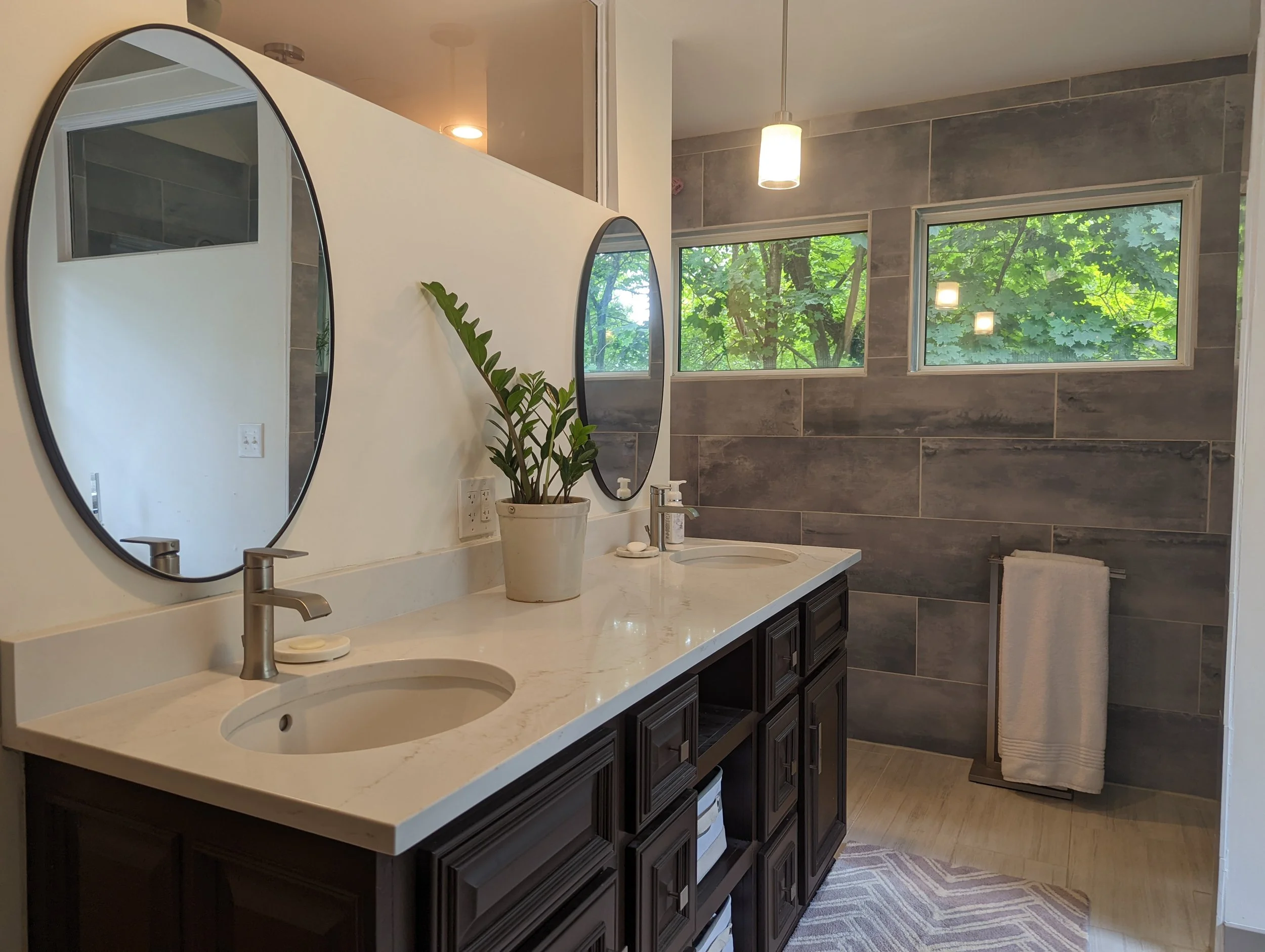 A bathroom with a white vanity, dark wood cabinets, two oval mirrors, a potted plant, and large windows showing green trees outside.