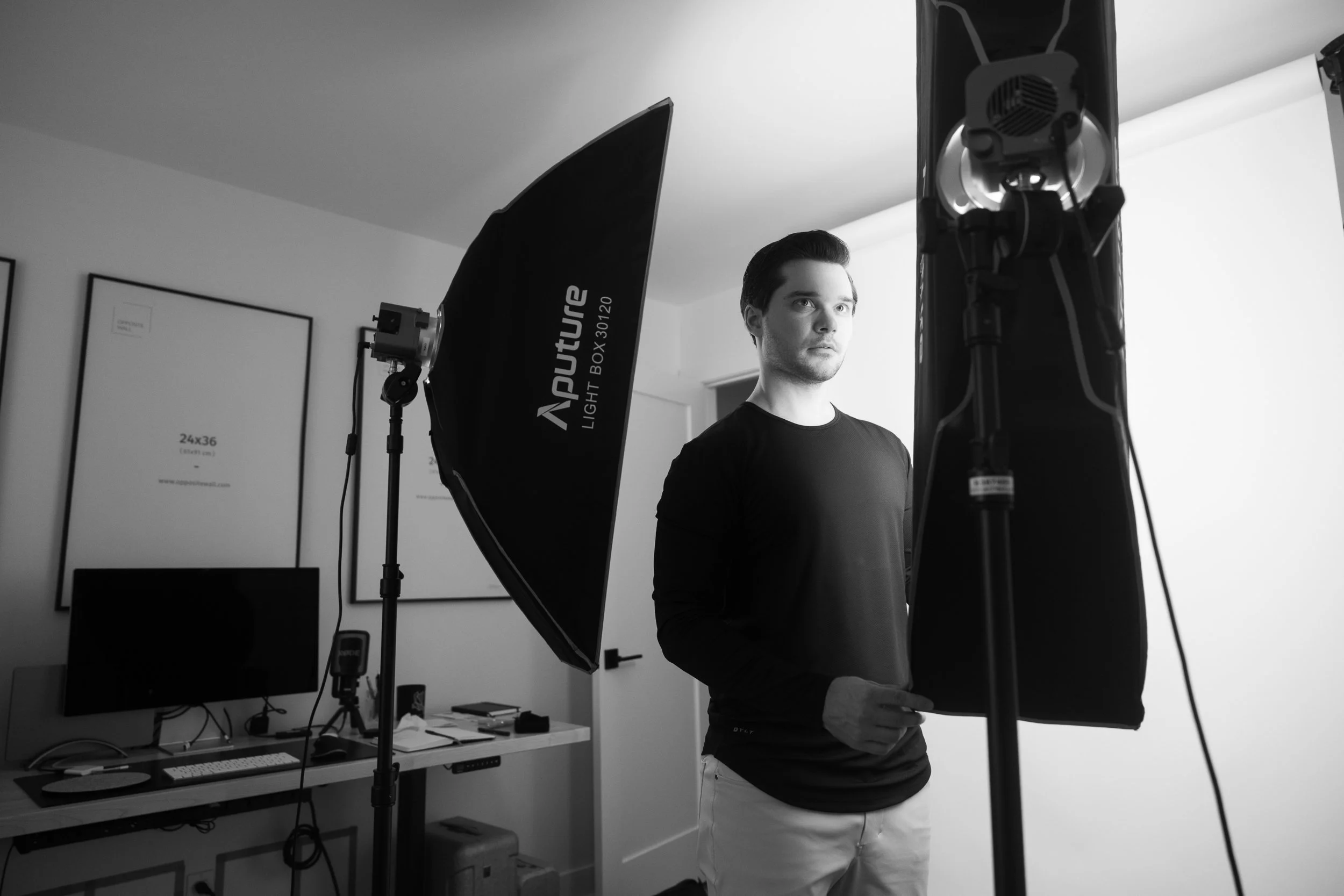 Black and white photo of a young man setting up studio lights in front of a white paper background.