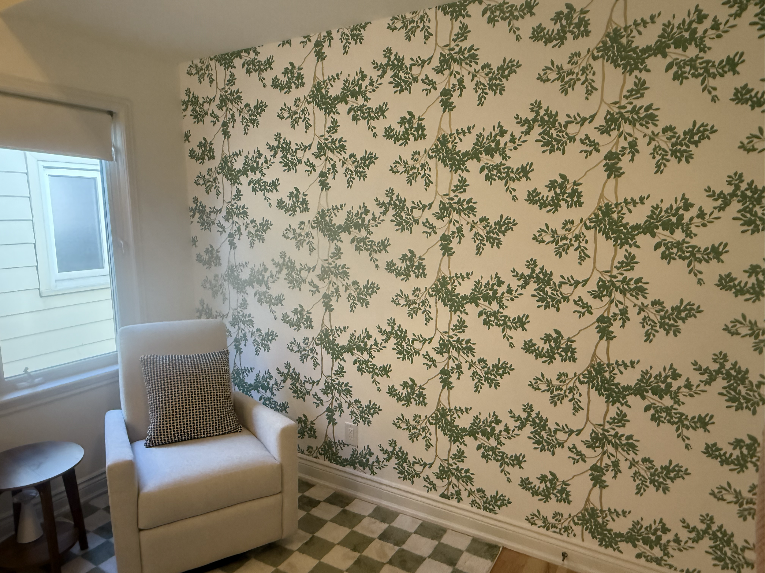 Living room with patterned wallpaper featuring green leaves, a beige armchair with a checkered pillow, a small side table, and a window with a closed white blind. Wallpaper. Interior House Painting. Heather Feather Painting and Design. Toronto.