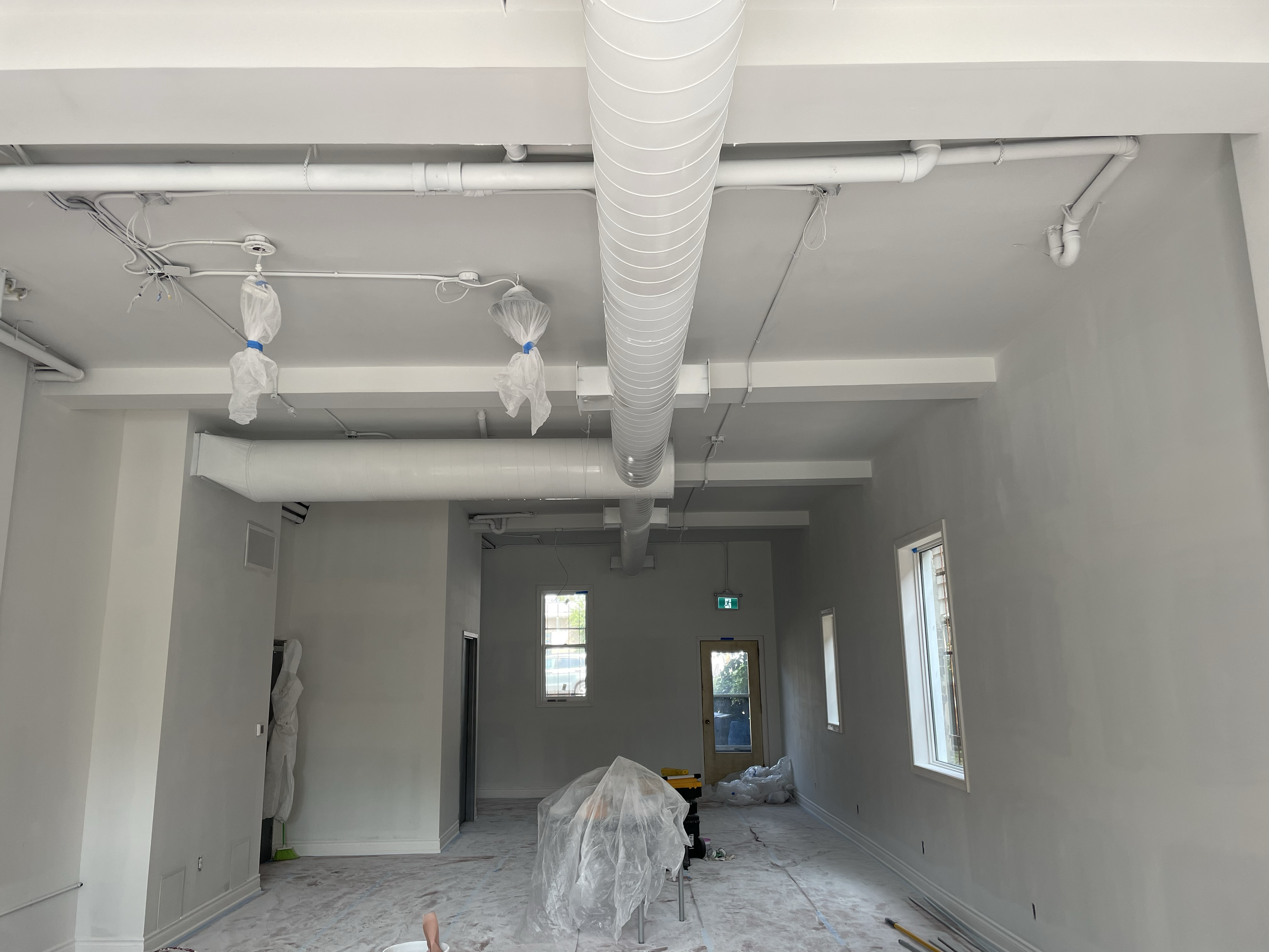 A room under construction with white walls and large windows. Exposed pipes run along the ceiling, and plastic sheeting covers the floor and some equipment. Commercial Painting. Heather Feather Painting and Design. Toronto. Spraying.