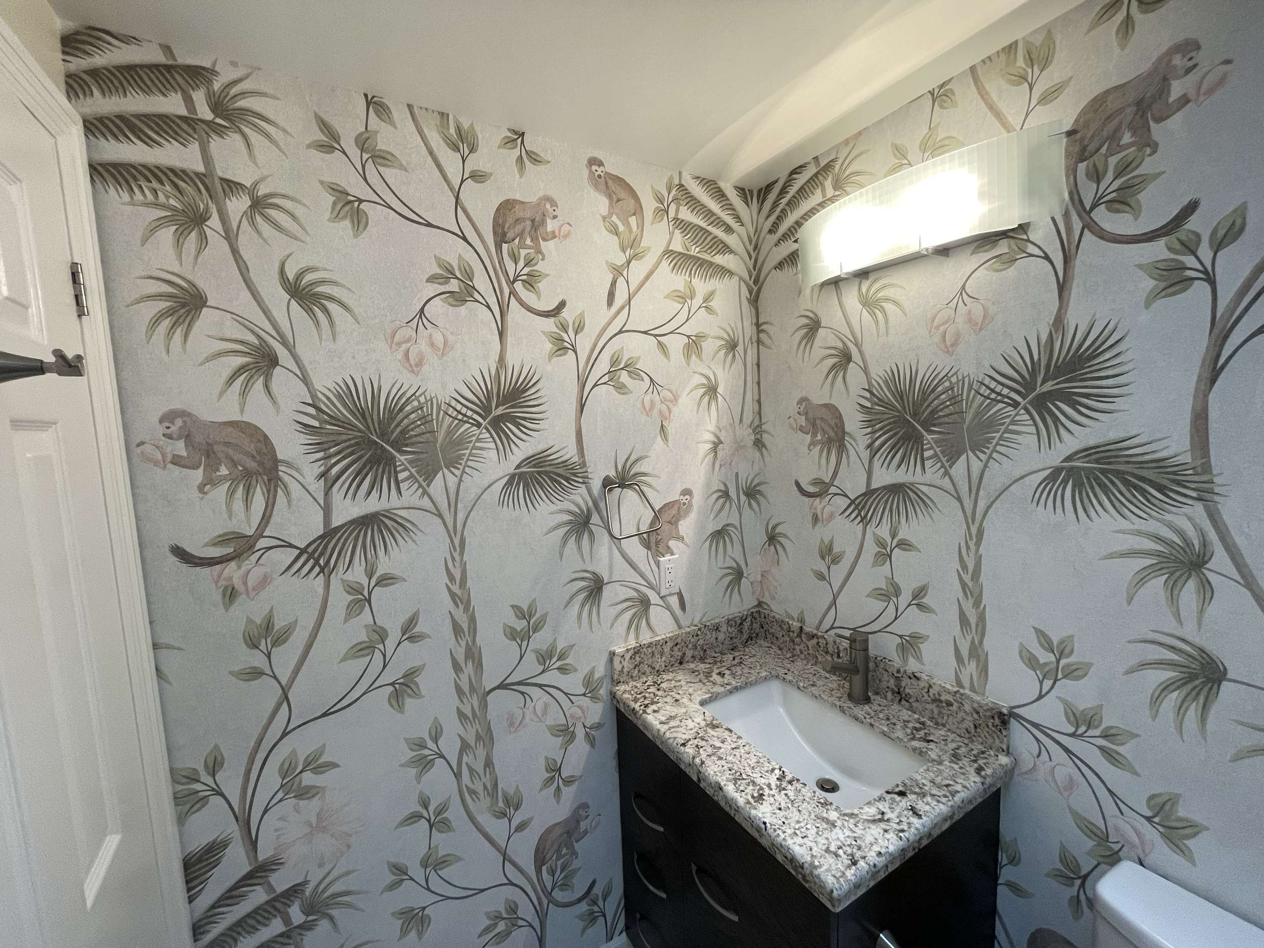 A bathroom with wallpaper featuring monkeys and tropical plants, a granite countertop with a white sink, and a window with a frosted glass cover. Interior House Painting. Heather Feather Painting and Design. Toronto.