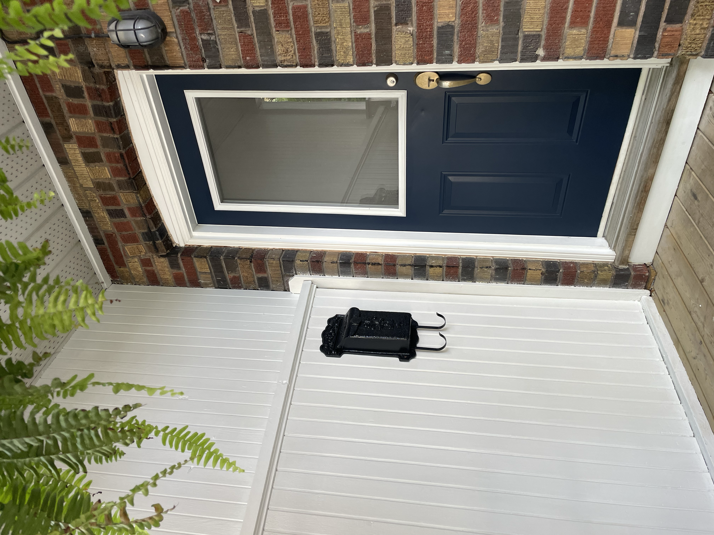 A front door painted dark blue with a gold handle and lock, set in a brick wall with a small glass window, and a porch area with a tiled floor and a black decorative lantern. Exterior House Painting. Heather Feather Painting and Design. Toronto.