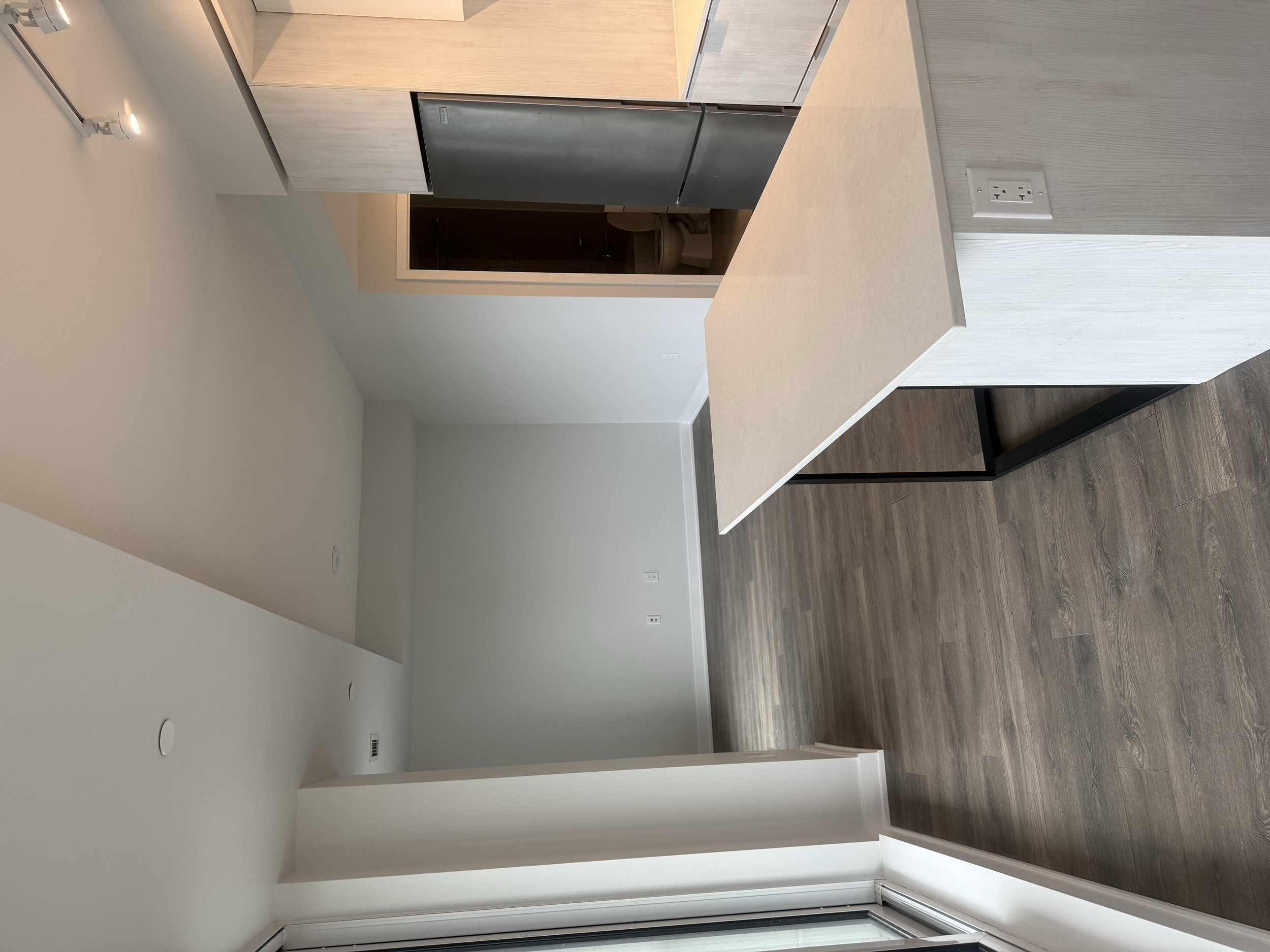 Empty kitchen with a beige countertop, wood cabinets, and stainless steel appliances, including a refrigerator. A doorway reveals a bathroom with a toilet inside. Interior House Painting. Heather Feather Painting and Design. Toronto. Condo