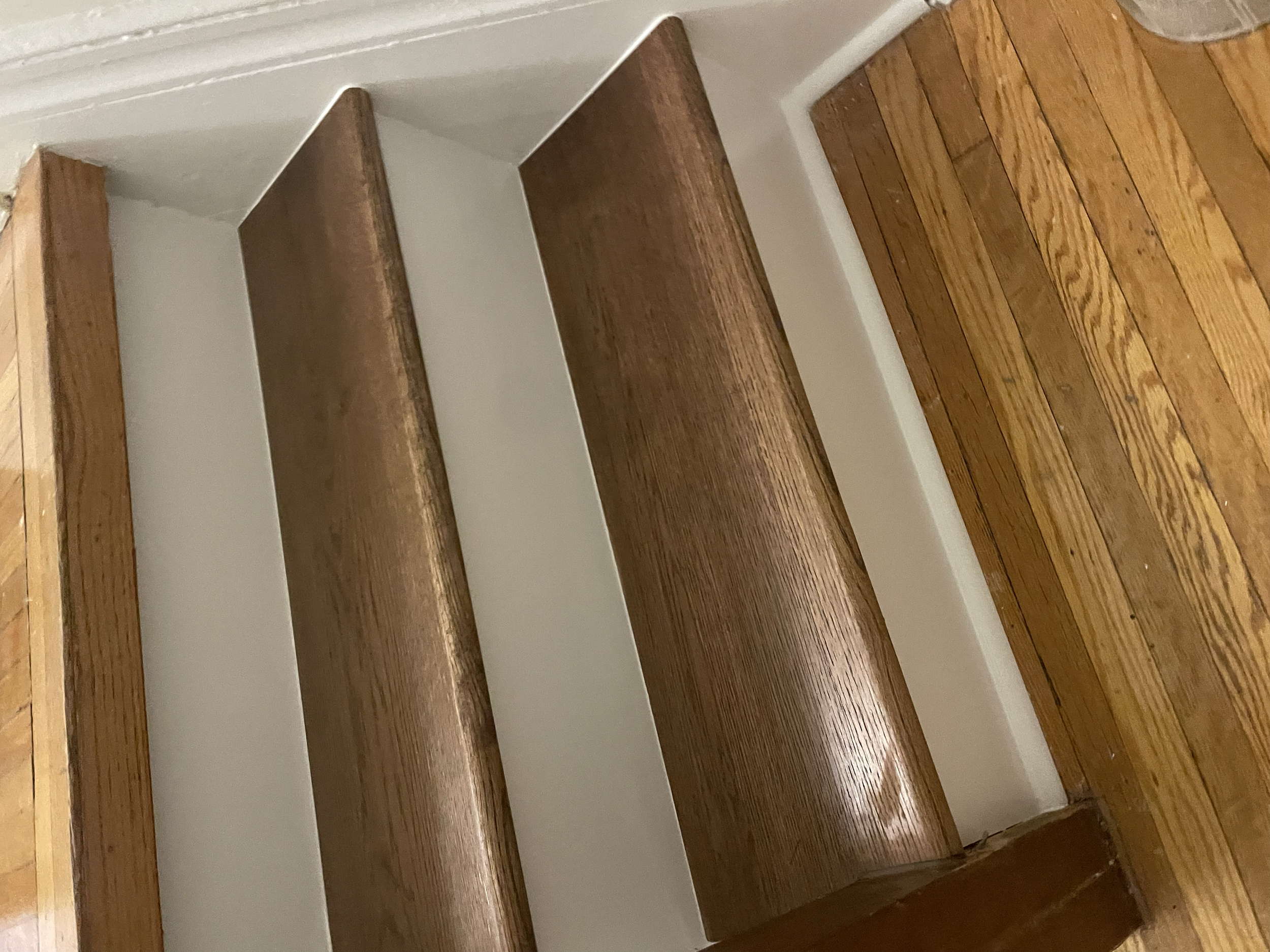 A wooden staircase with three steps, against a white wall, with a hardwood floor at the bottom. Interior House Painting. Heather Feather Painting and Design. Toronto. Stair Refinishing.