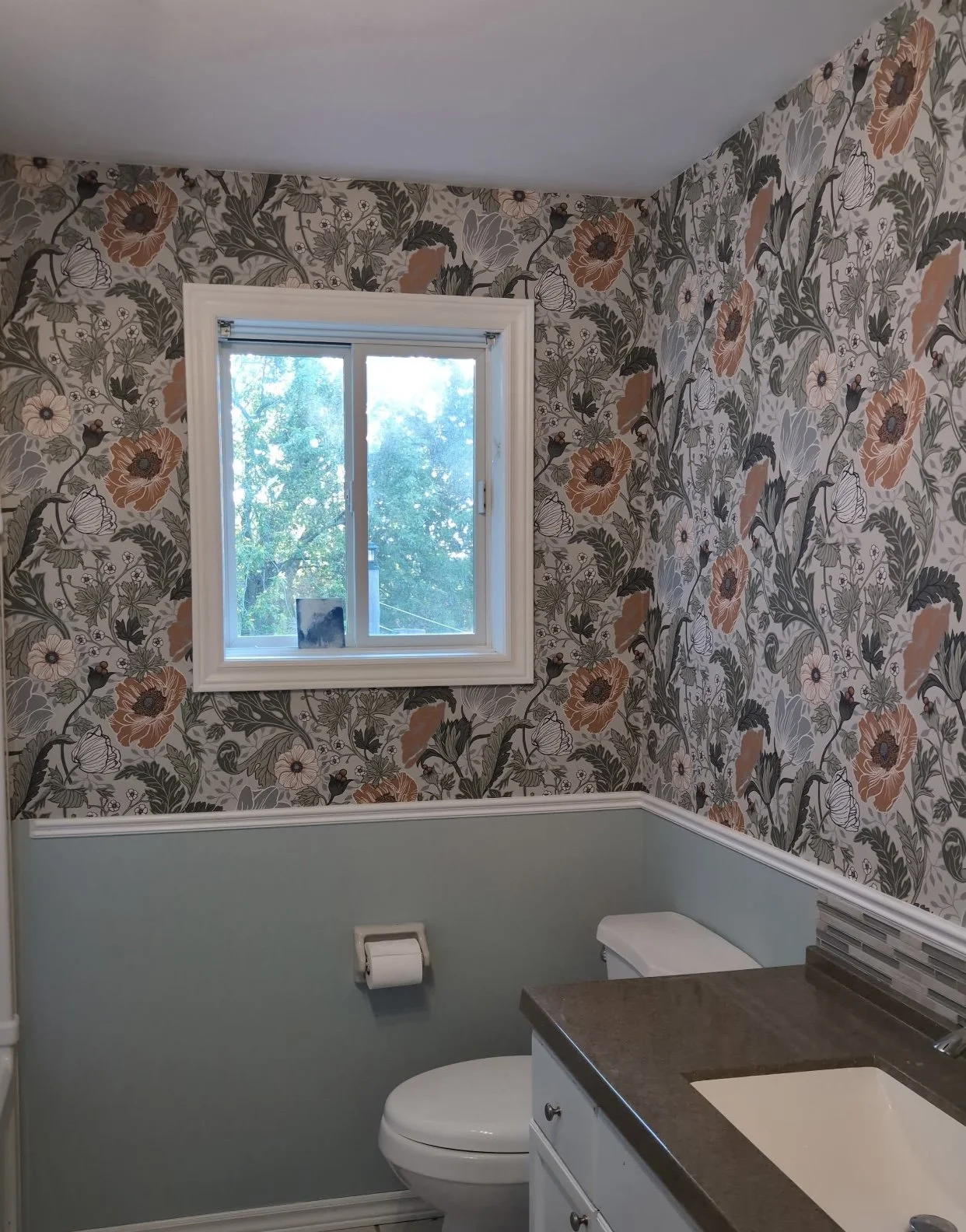 A bathroom with floral wallpaper, a small window showing trees outside, a toilet, and a bathroom sink with a gray countertop.
Wallpaper. Interior House Painting. Heather Feather Painting and Design. Toronto.