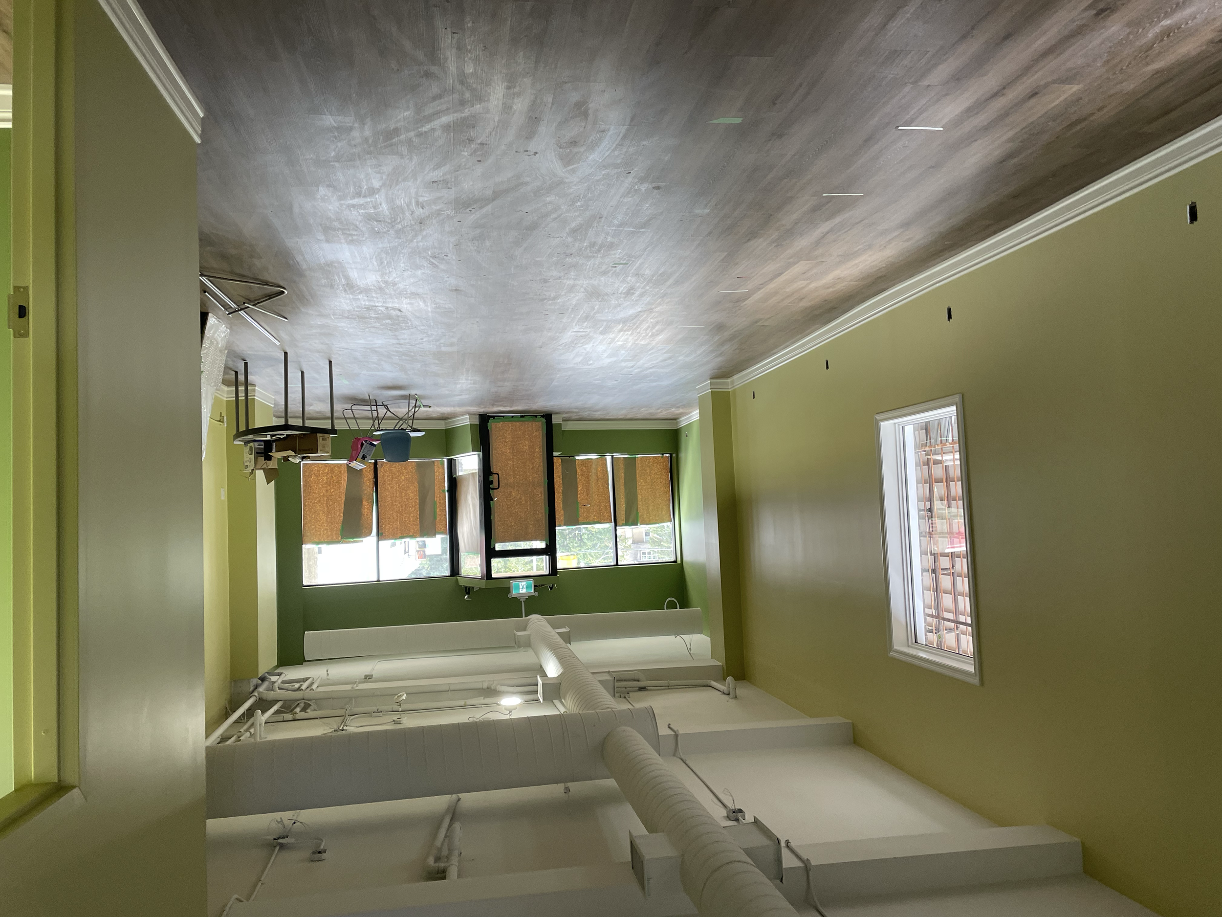 An open room with bright green walls, two windows, and a green door, with construction materials and equipment visible, including a large white pipe and a ladder. Commercial Painting. Heather Feather Painting and Design. Toronto. 