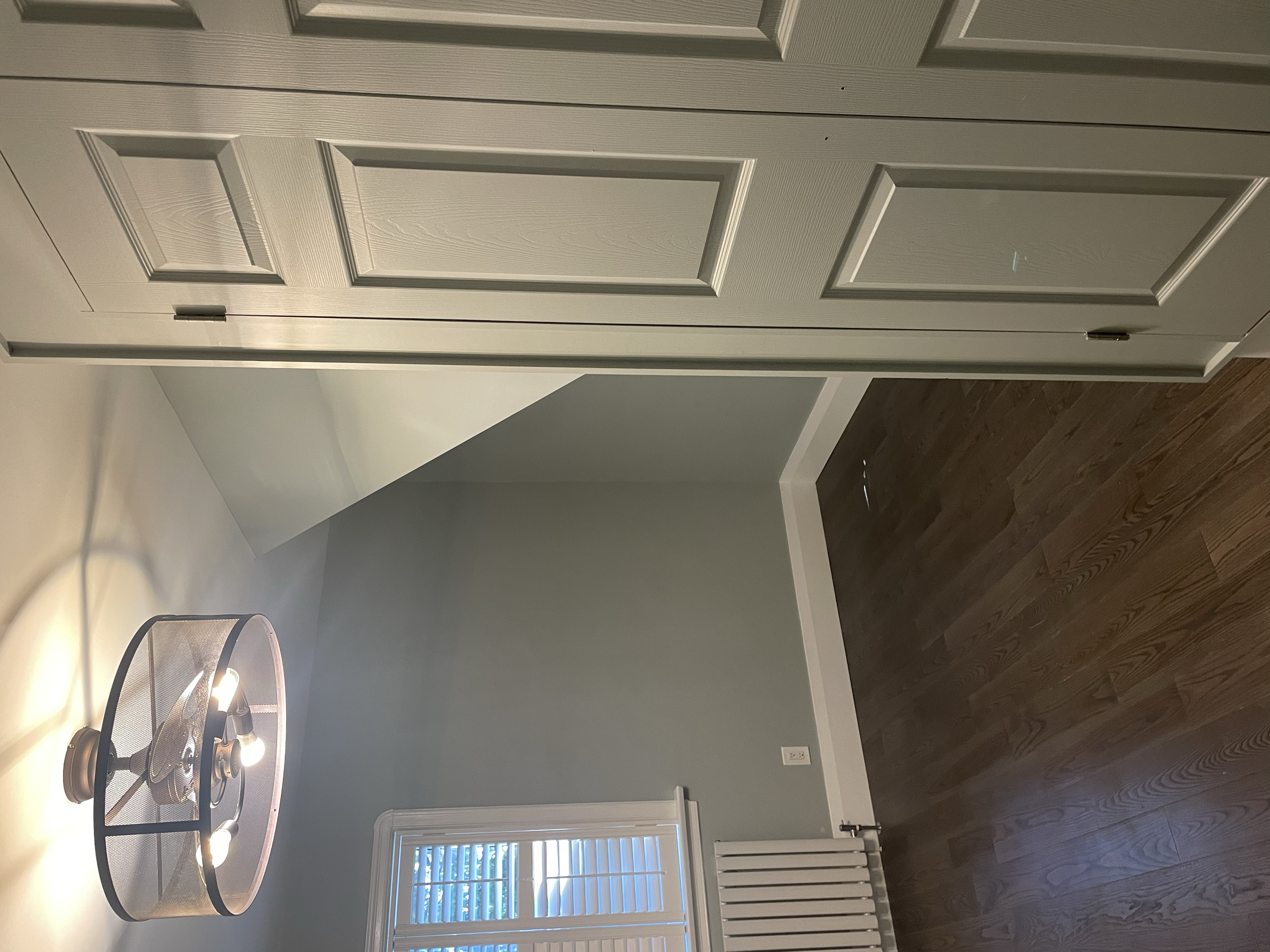 Interior room with a dark wooden floor, a white heater below a window with blinds, gray painted walls, a light fixture with multiple bulbs, and a corner of a white door. Interior House Painting. Heather Feather Painting and Design. Toronto.