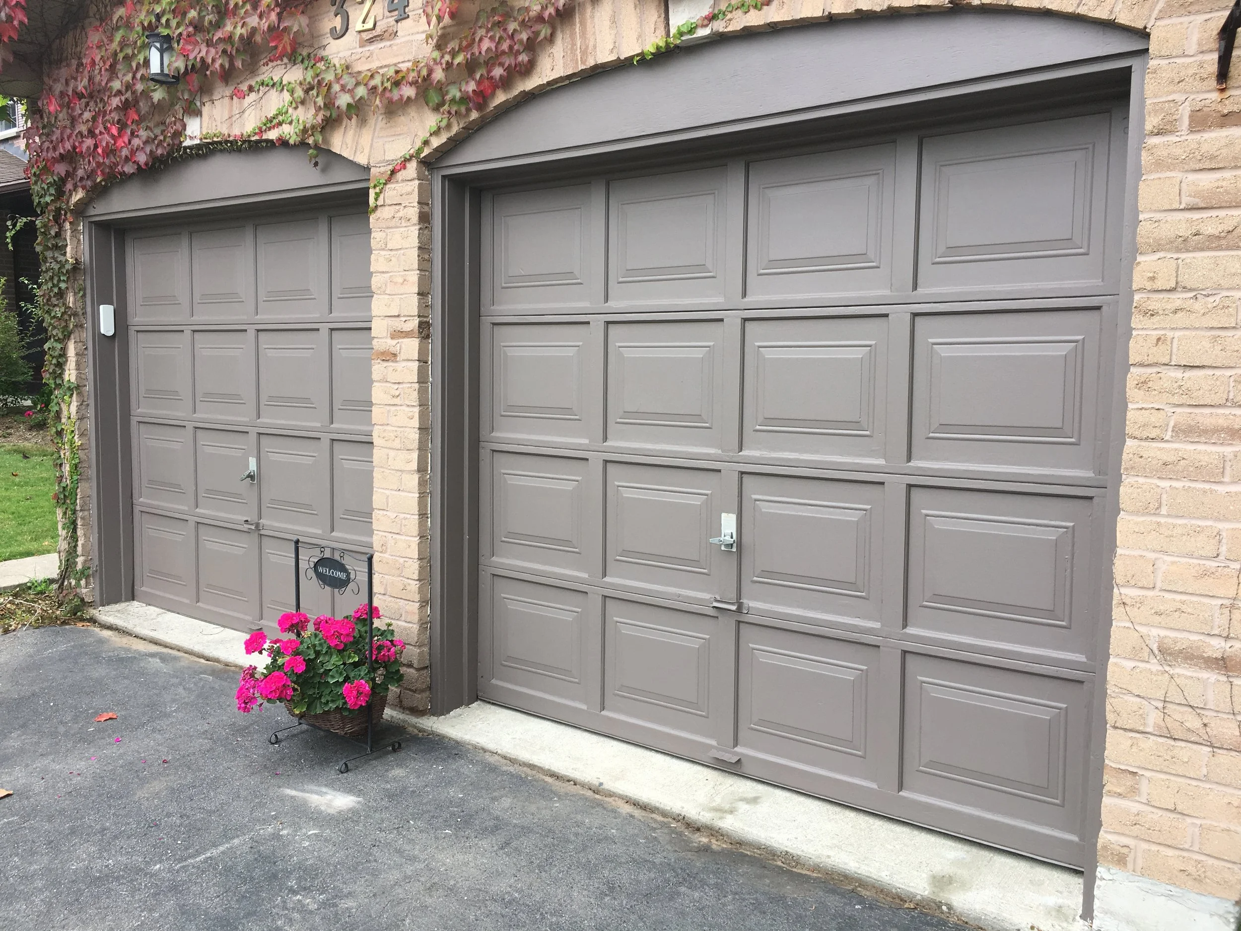 Two gray garage doors with brick walls on each side, potted pink flowers, and a black welcome sign in front of the left door.
Exterior House Painting. Heather Feather Painting and Design. Toronto. Garage Door.