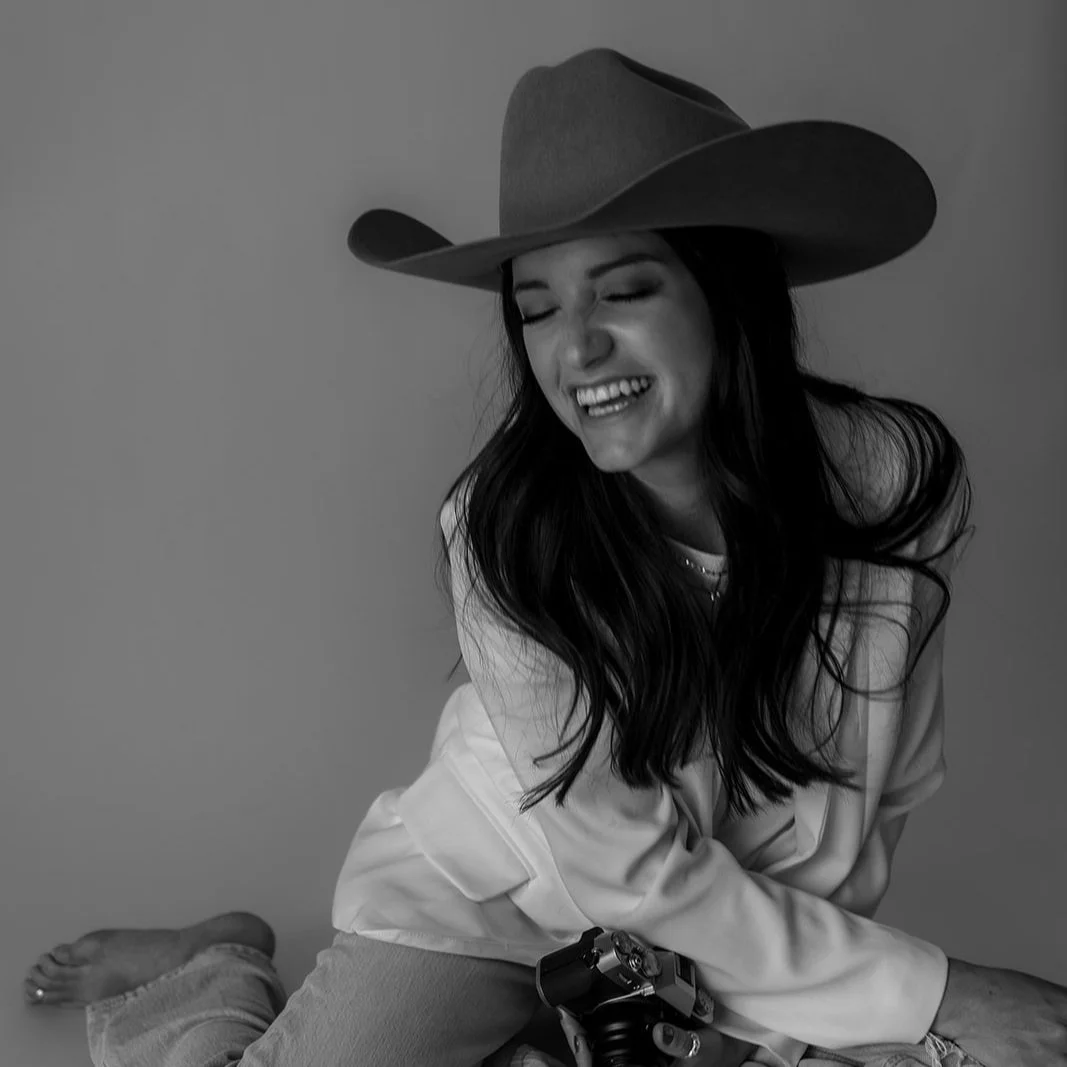 Black and white photo of a Jessica Luann wearing a cowboy hat, laughing, and sitting with a camera.