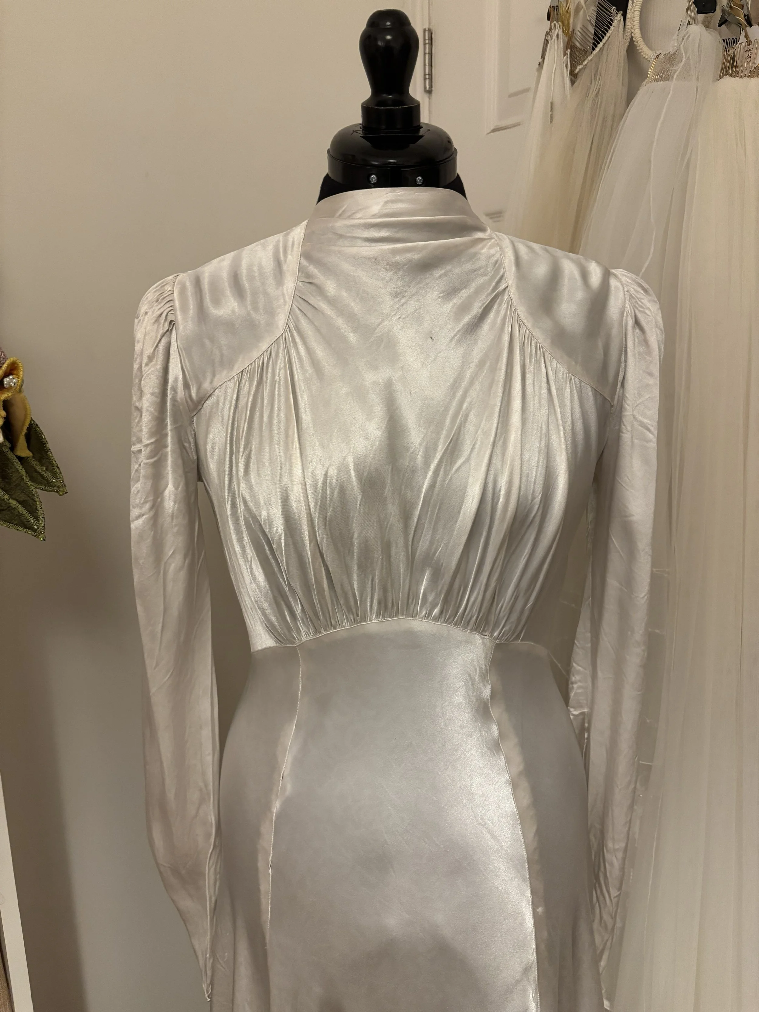 Vintage cream-colored satin dress with long sleeves, a high collar, pleated bodice, and fitted waist, displayed on a mannequin.