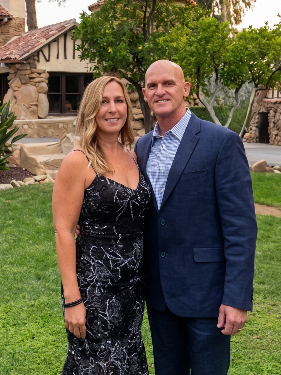 A man and woman standing together on a grassy lawn in front of a stone house with lush trees.
