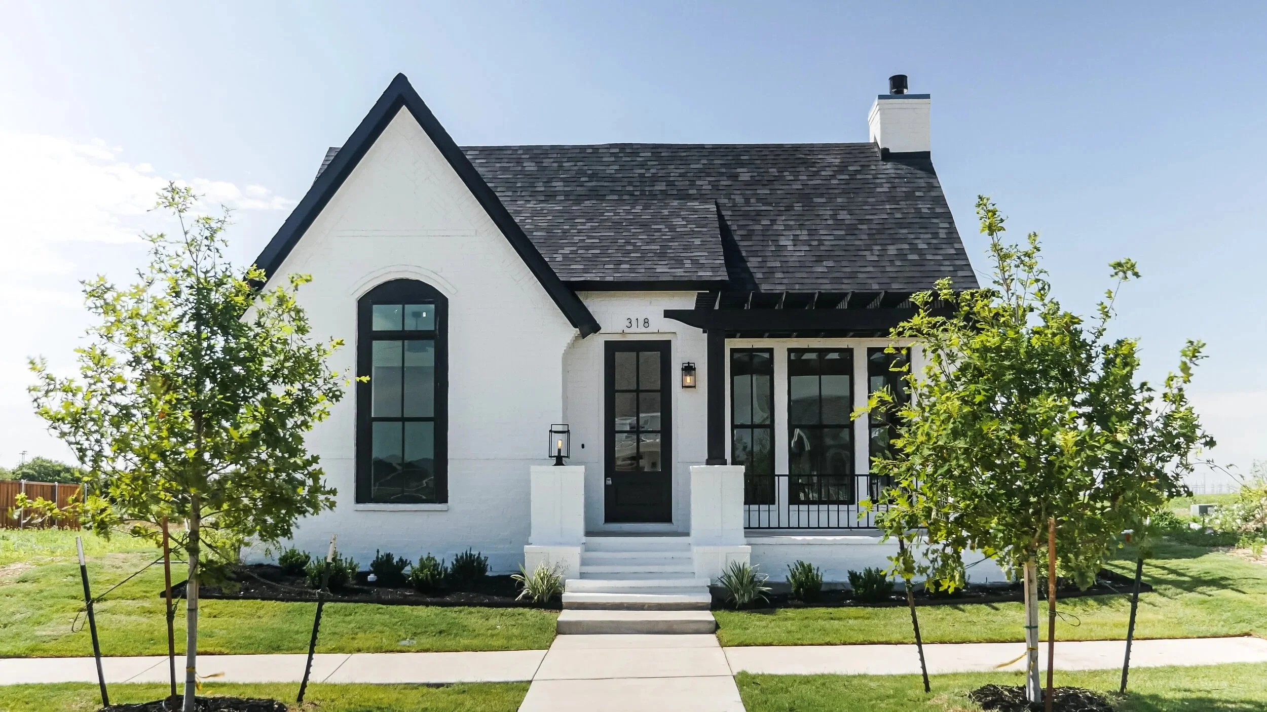 A modern white house with black trim and large windows, surrounded by trees and a lawn, under a clear blue sky.