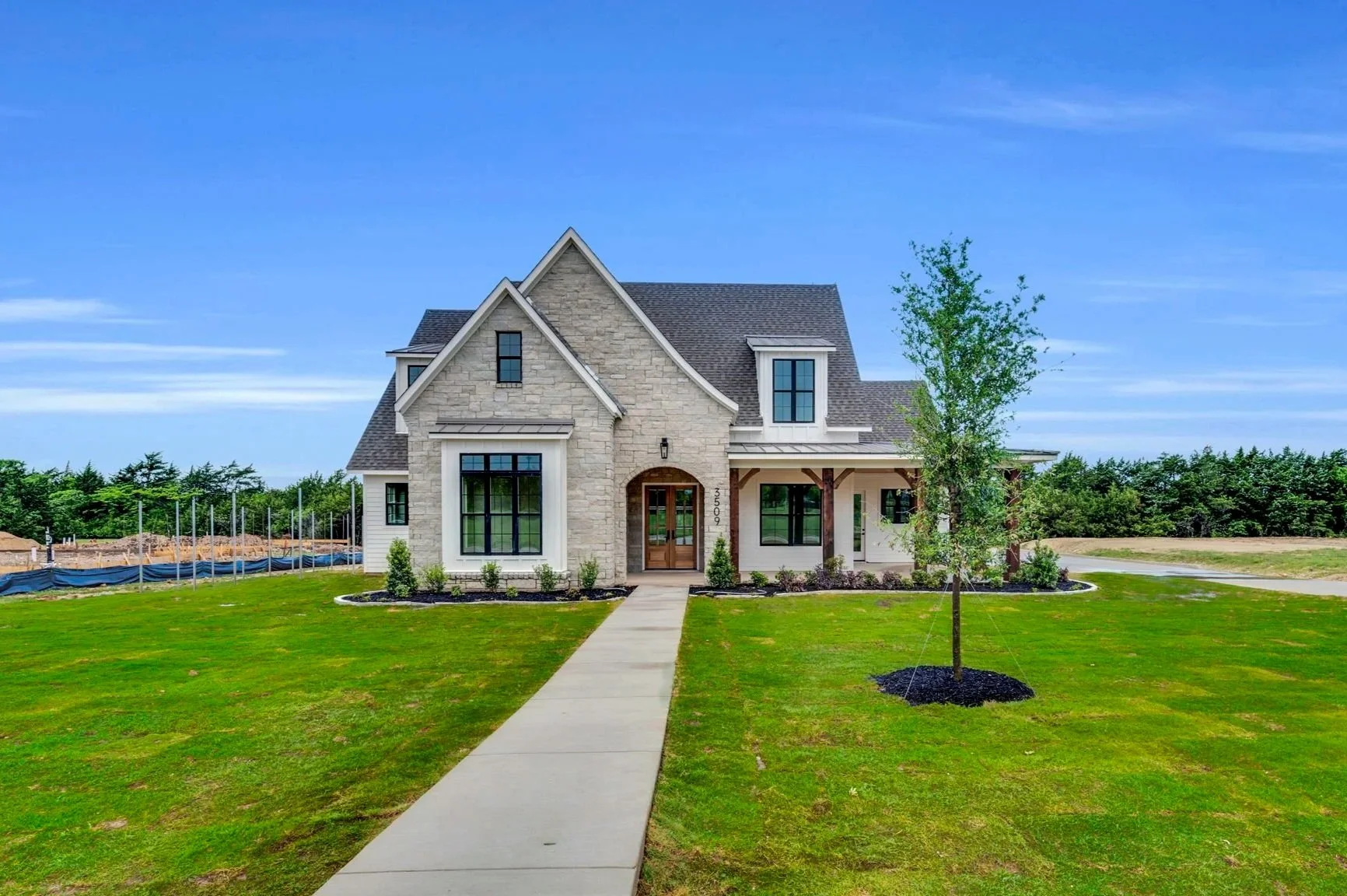A newly built two-story house with a stone facade, large windows, and a front porch, surrounded by green lawn and small trees under a blue sky.