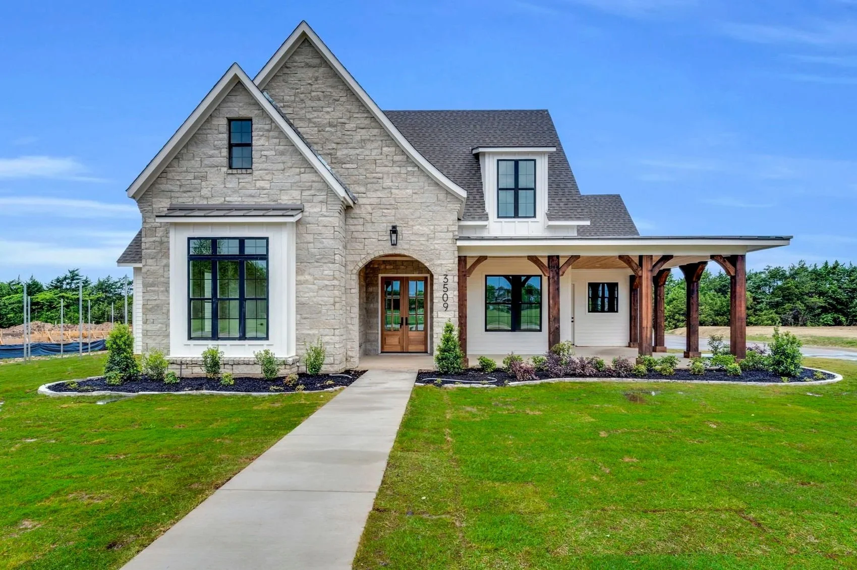 Front view of a modern house with stone and siding exterior, large front window, wooden front door, and a covered porch supported by wooden beams. The house is numbered 3509 and has a well-maintained lawn with small shrubs and mulch beds.