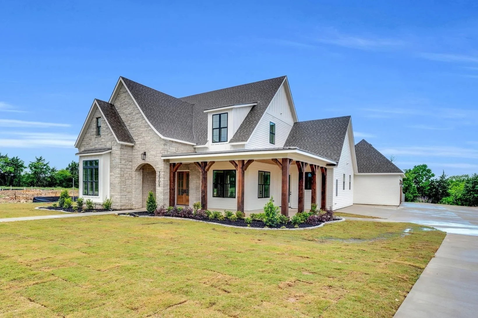 New two-story house with a mix of stone and white siding, front porch with wooden supports, large front yard, and driveway on the right, under a clear blue sky.