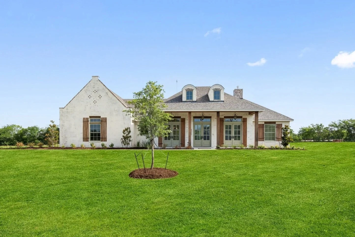 Front view of a large house with white brick exterior, multiple windows with shutters, a front porch with columns, and a well-maintained green lawn with a young tree in the foreground.
