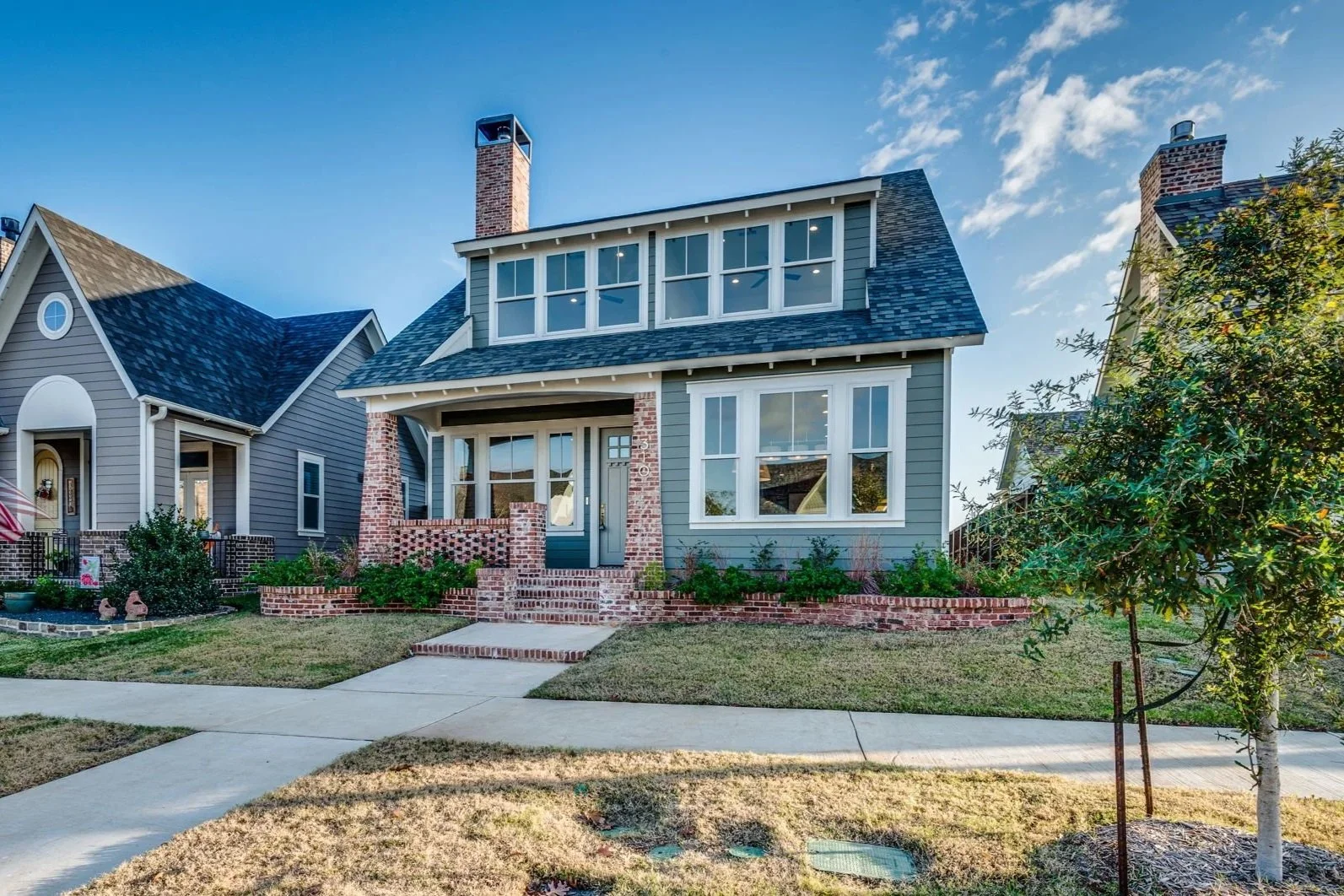 A two-story house with gray siding and brick accents, large front windows, a small front porch, and a manicured lawn.