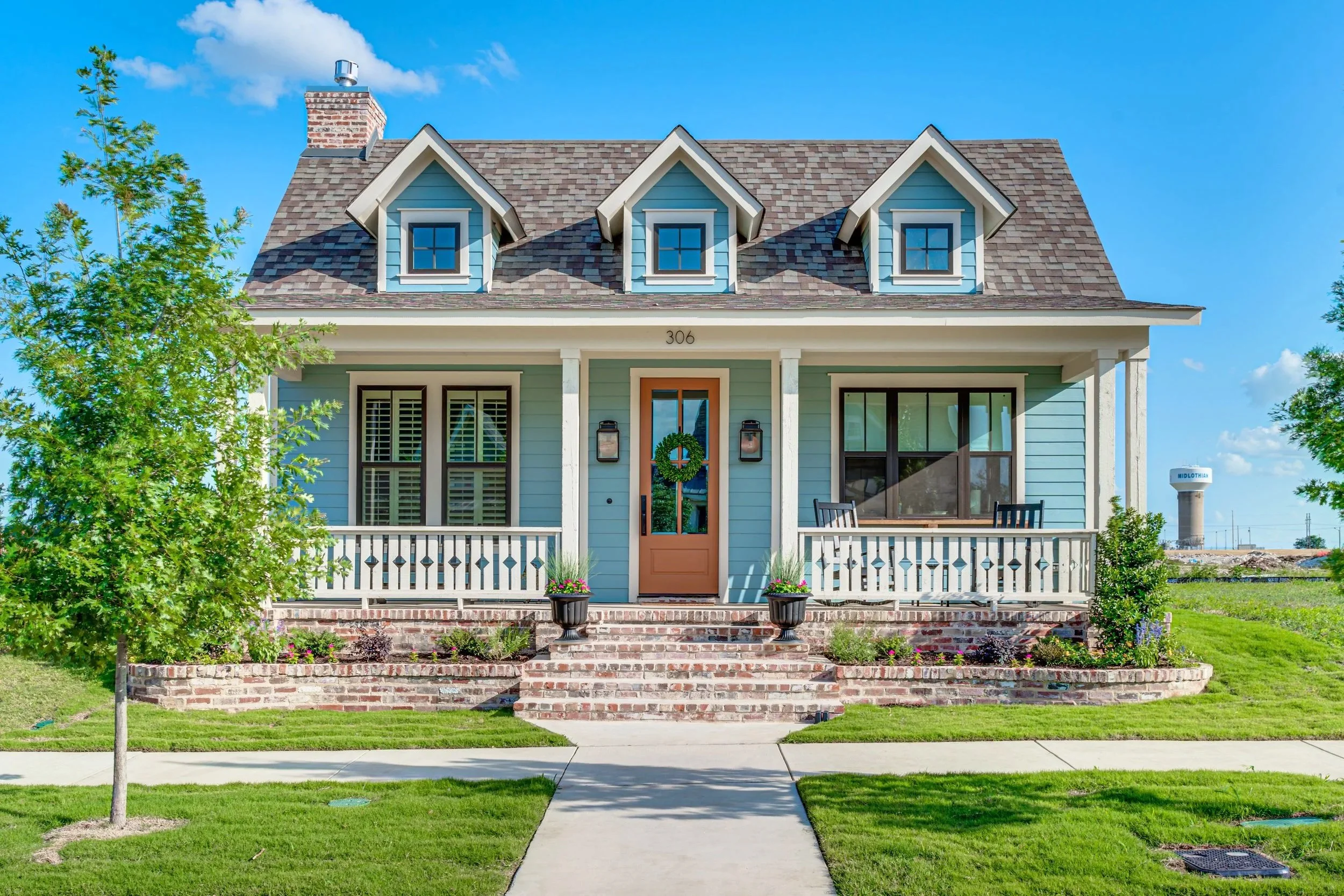 Front view of a light blue house with a porch, three dormer windows on the roof, a brick chimney, and a green lawn with a sidewalk and small trees.