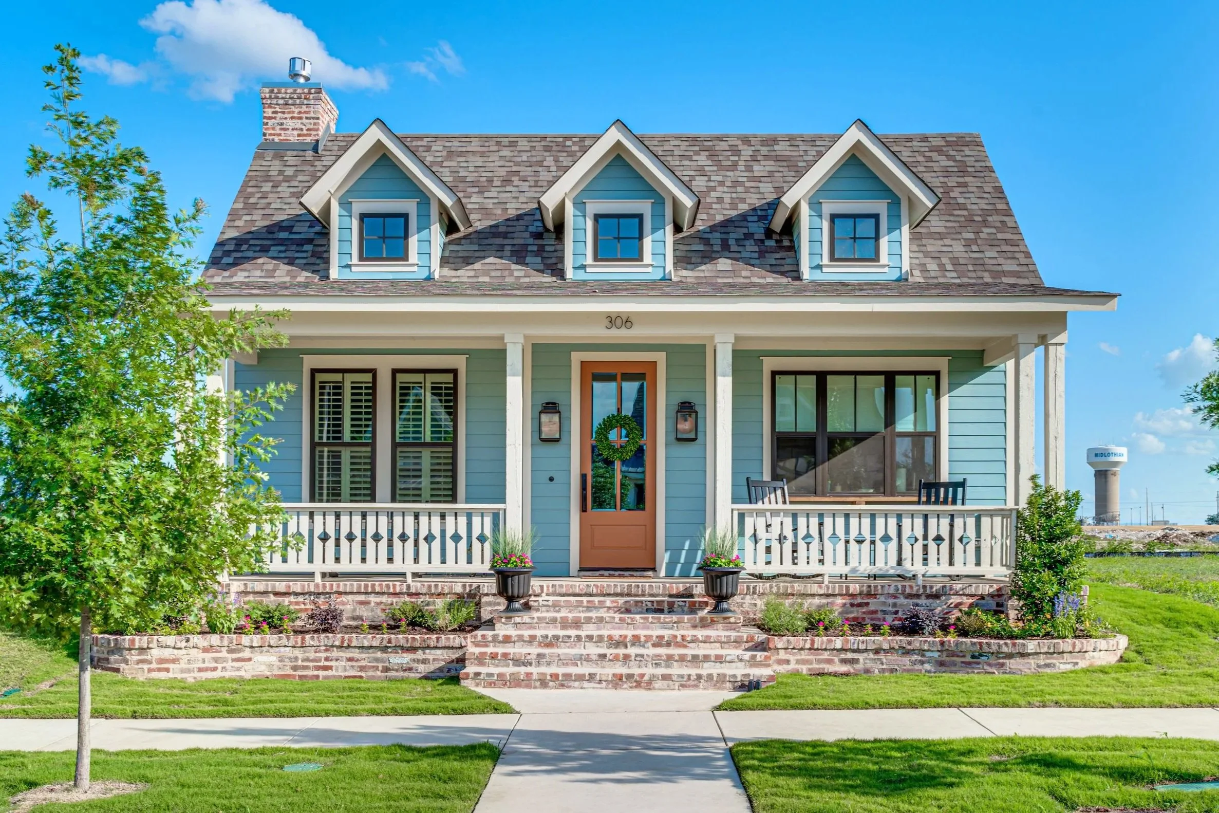 Front view of a light blue two-story house with a porch, three dormer windows on the roof, a brick chimney, and a front yard with a sidewalk, small tree, and flower beds.