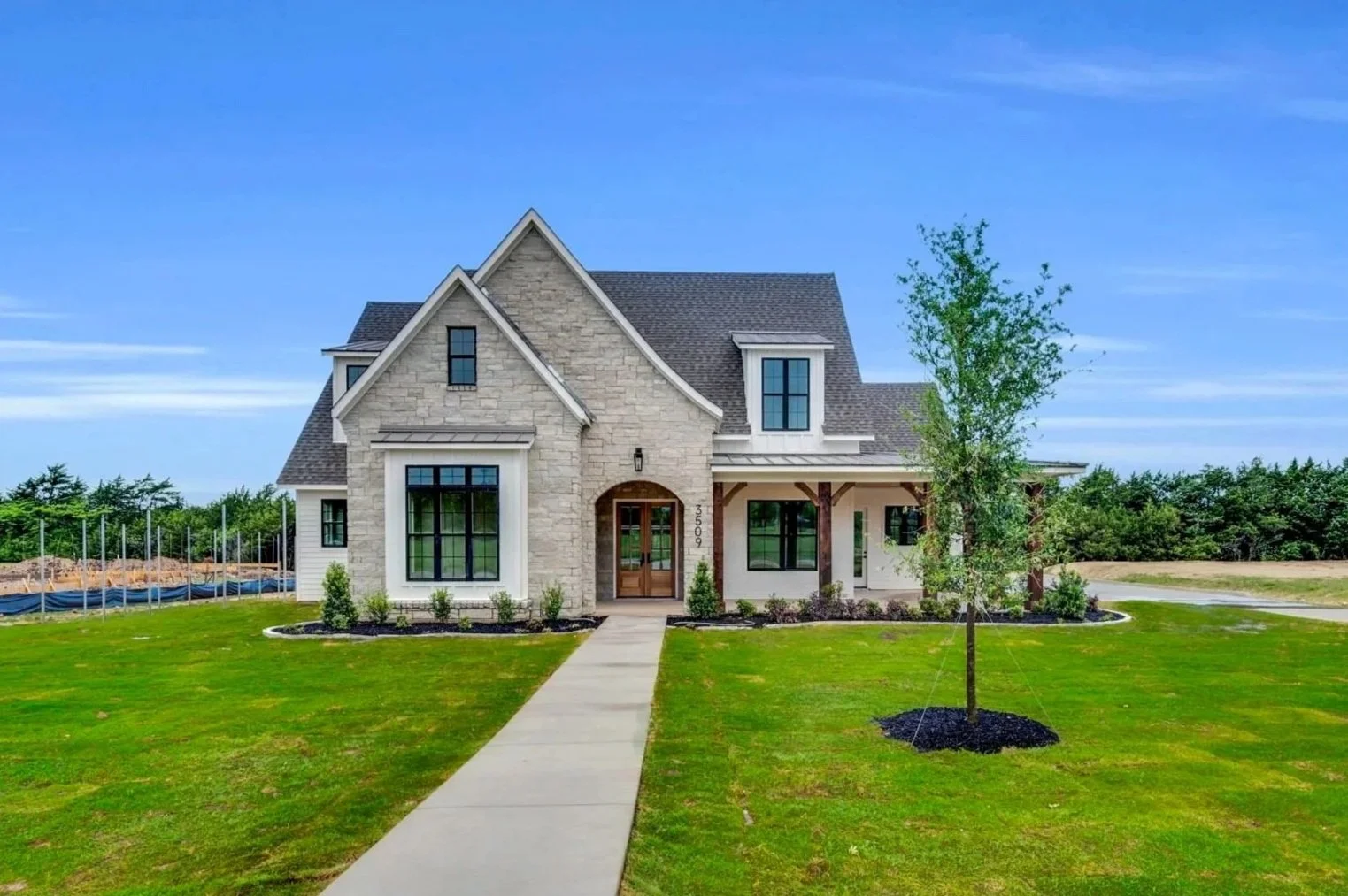 Newly built two-story house with stone and white siding, large front yard with lush green grass, small trees, and a paved walkway leading to the front door, under a blue sky with some clouds.