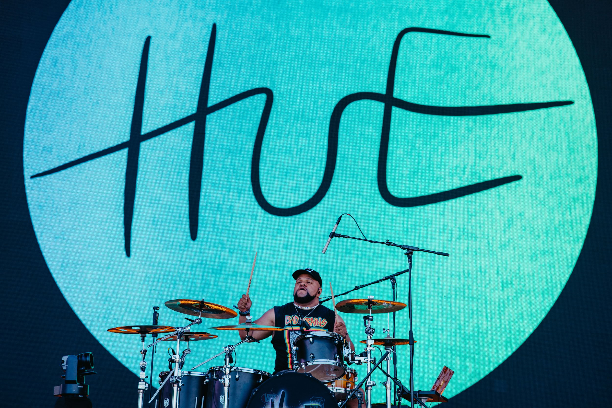 Musician playing drums on stage, with a large screen behind featuring the word 'Life' in stylized script.