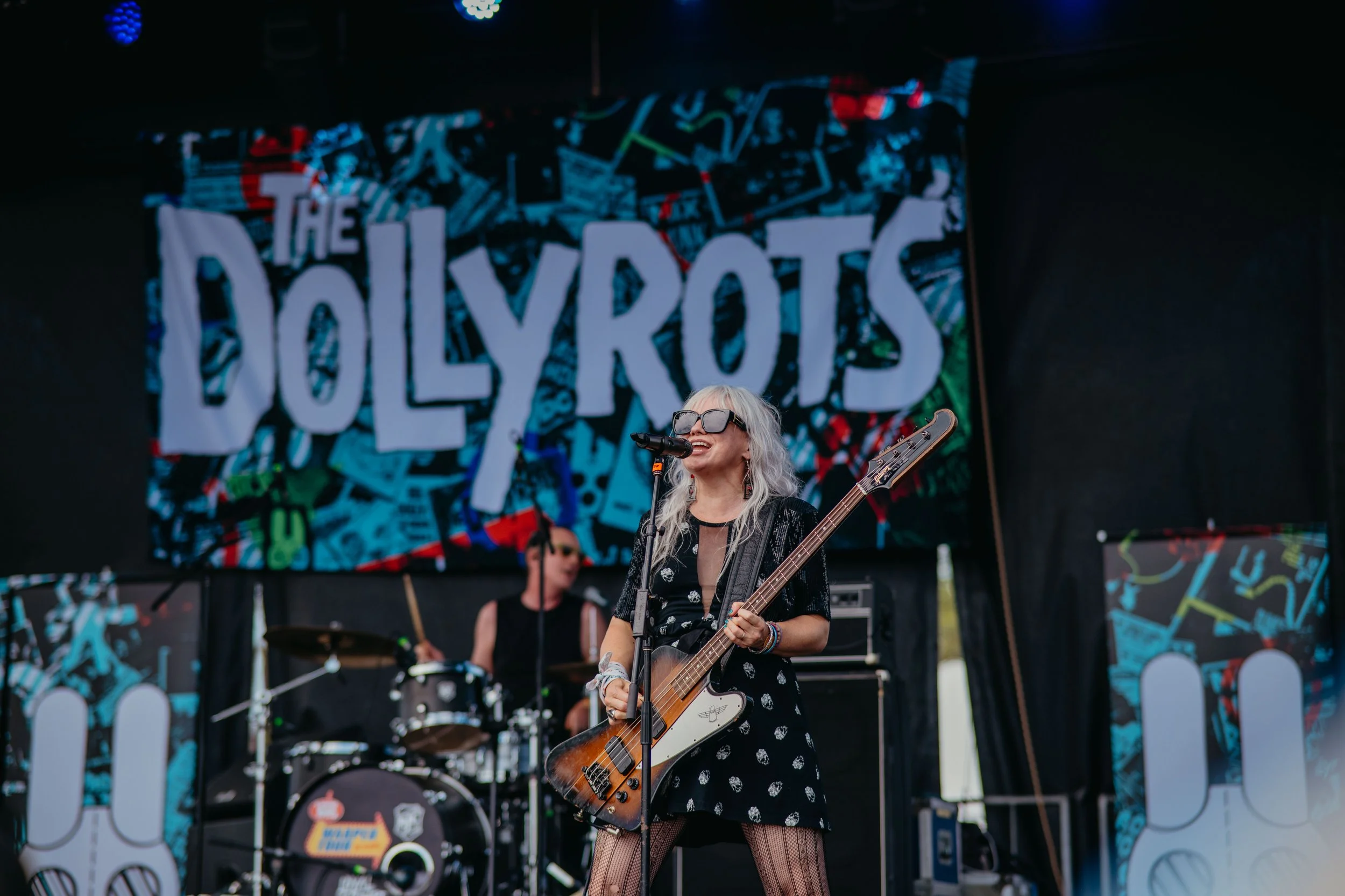 A female musician with gray hair and sunglasses singing into a microphone while playing an electric guitar during a live outdoor performance. Behind her, a drummer is visible, and a colorful banner with the words 'The POLYVOTS' is displayed.