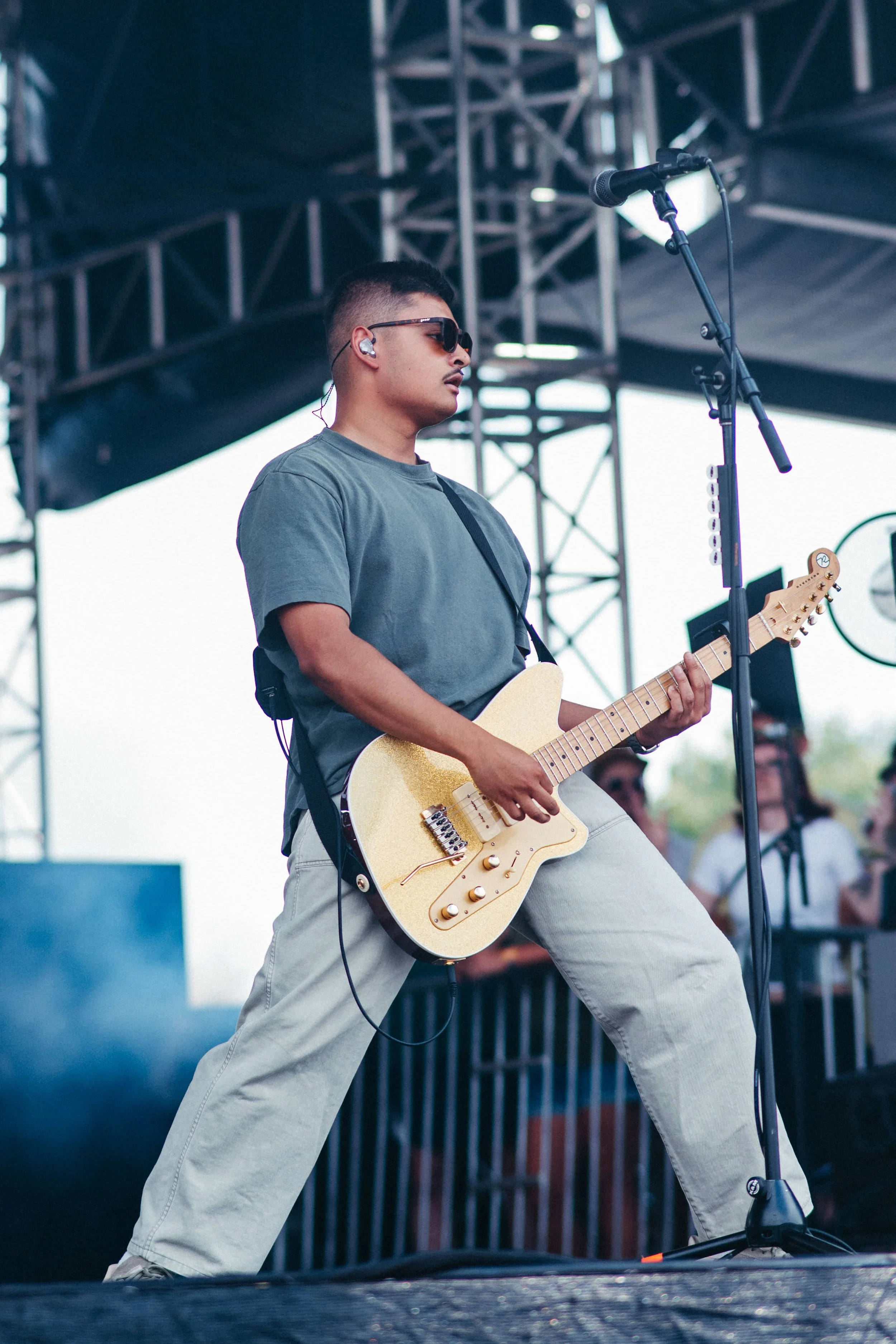 A male musician playing an electric guitar on stage during an outdoor concert. He wears sunglasses, a gray t-shirt, and beige pants, with stage equipment and a microphone in front of him.