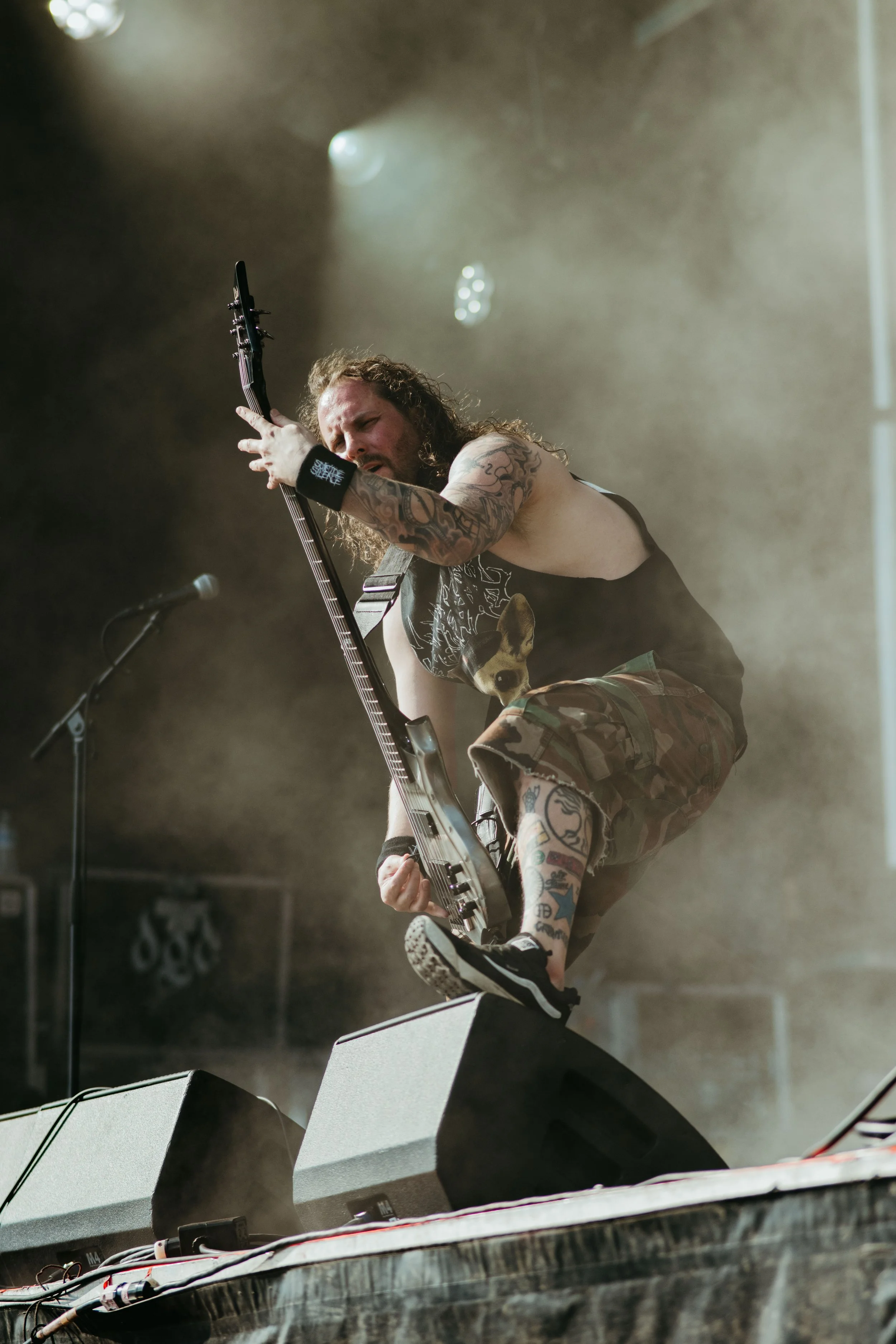 A shirtless musician with tattoos, long curly hair, and a wristband is playing an electric bass guitar on stage during a concert, with stage lights and fog creating an intense atmosphere.