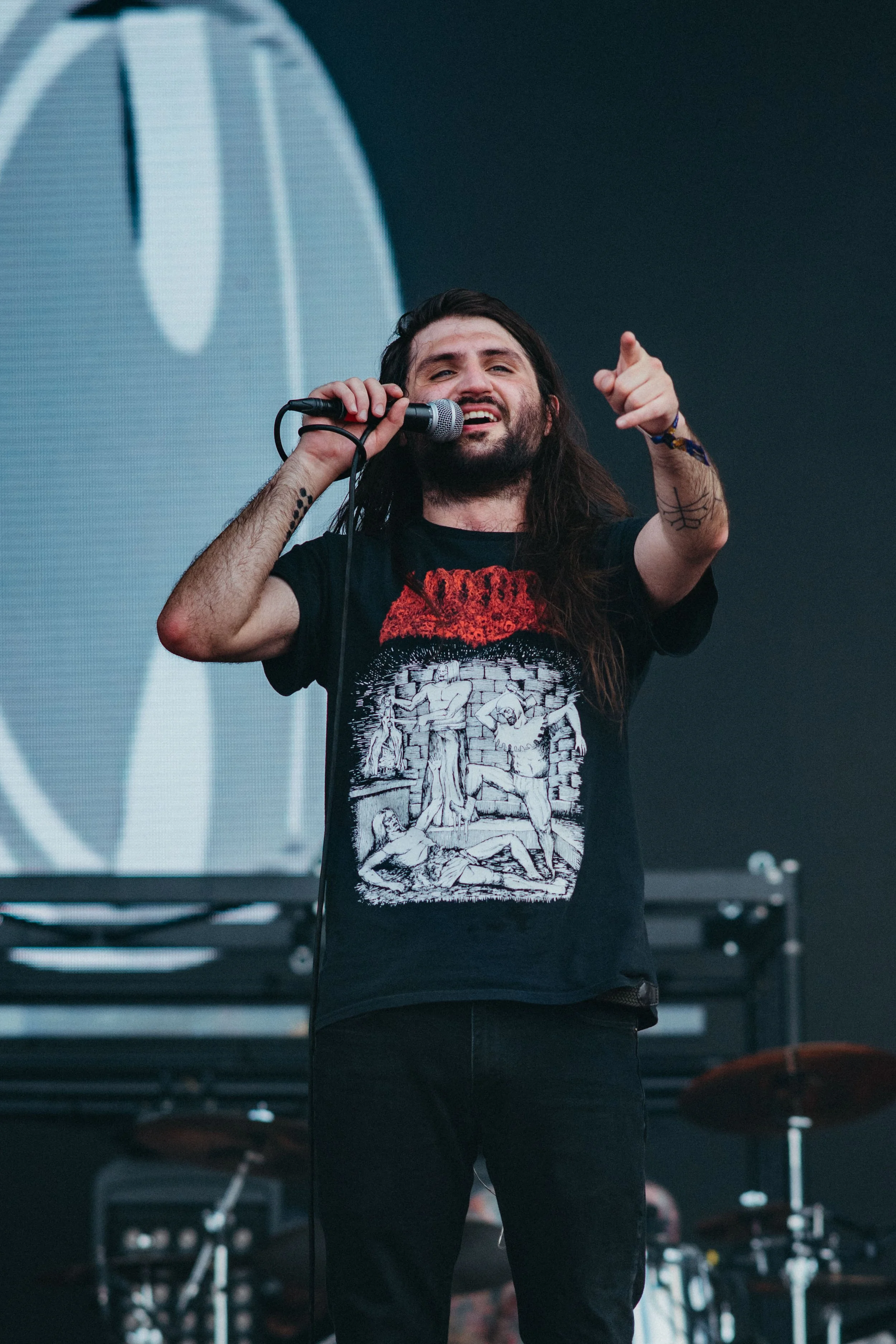 Male singer with long hair and beard performing on stage, holding a microphone, pointing towards the camera, wearing a black T-shirt with graphic art, standing in front of a digital screen background.