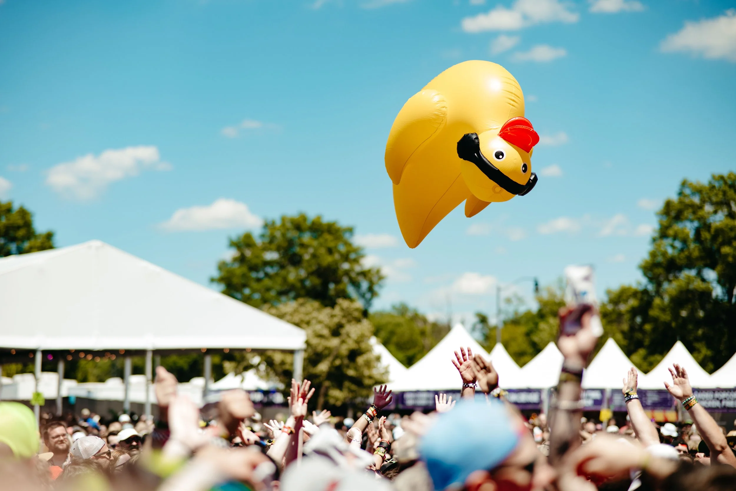 A yellow helium balloon shaped like a duck with a red crest, black beak, and large eyes, floating above a crowd at an outdoor event on a sunny day.