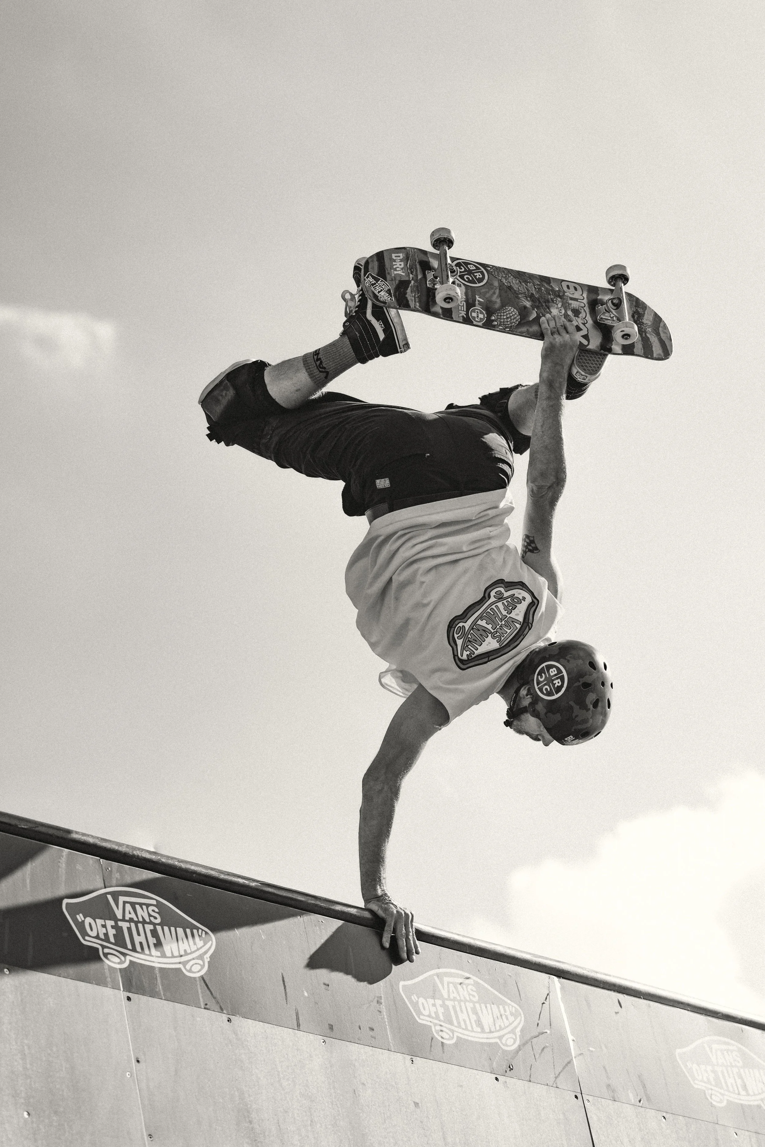 A skateboarder performing a handstand on the edge of a skate ramp, wearing a helmet and protective gear, with a "Vans Off The Wall" logo visible on the ramp.