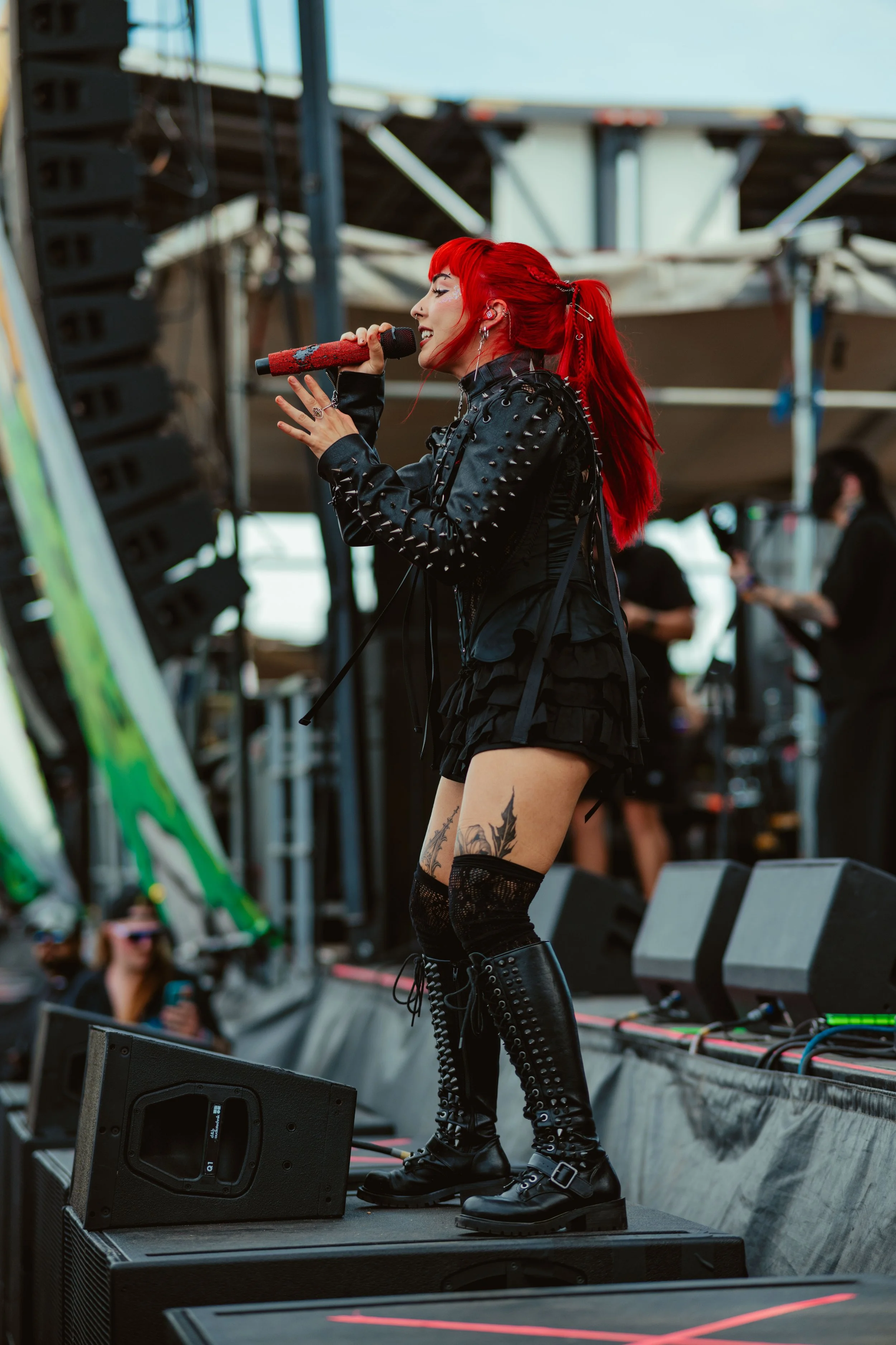 A woman with bright red hair singing into a microphone on stage, wearing a black leather jacket with spikes, a black dress, fishnet stockings, and tall black boots. There are large speakers on stage and people in the background.