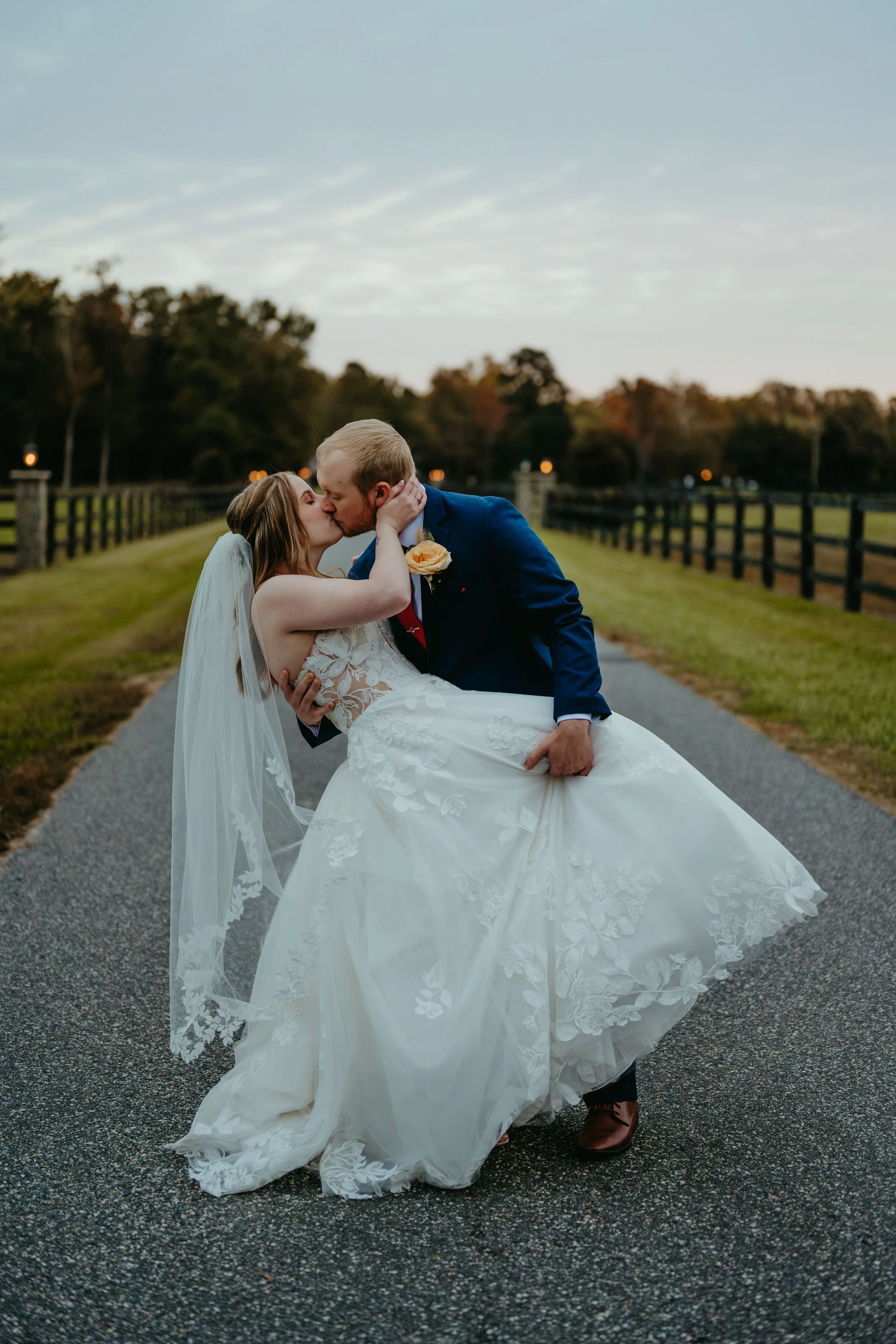 A bride and groom share a kiss outdoors on a gray paved path, with the groom lifting the bride in his arms. The bride is wearing a lace wedding gown and veil, and the groom is dressed in a blue suit with a yellow boutonniere. Fall trees and a fence l