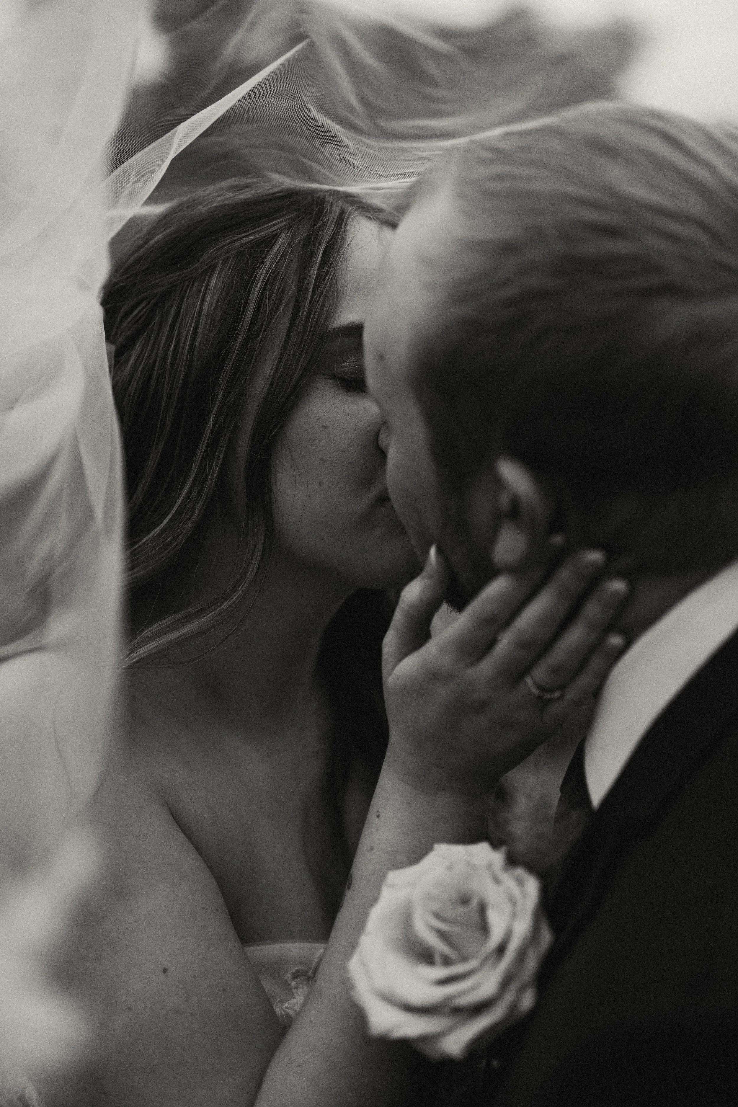 A black-and-white photograph of a bride and groom kissing during their wedding, with the bride wearing a veil and a corsage with a flower on her wrist.