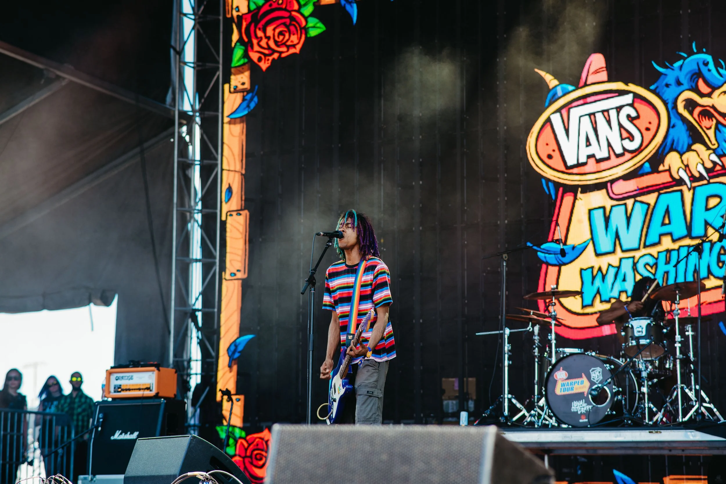 A musician performing on a stage with a colorful backdrop including the Vans logo and other graphic designs. The musician has multicolored hair, wears a striped shirt, and plays an electric guitar. He is in the band The Paradox.