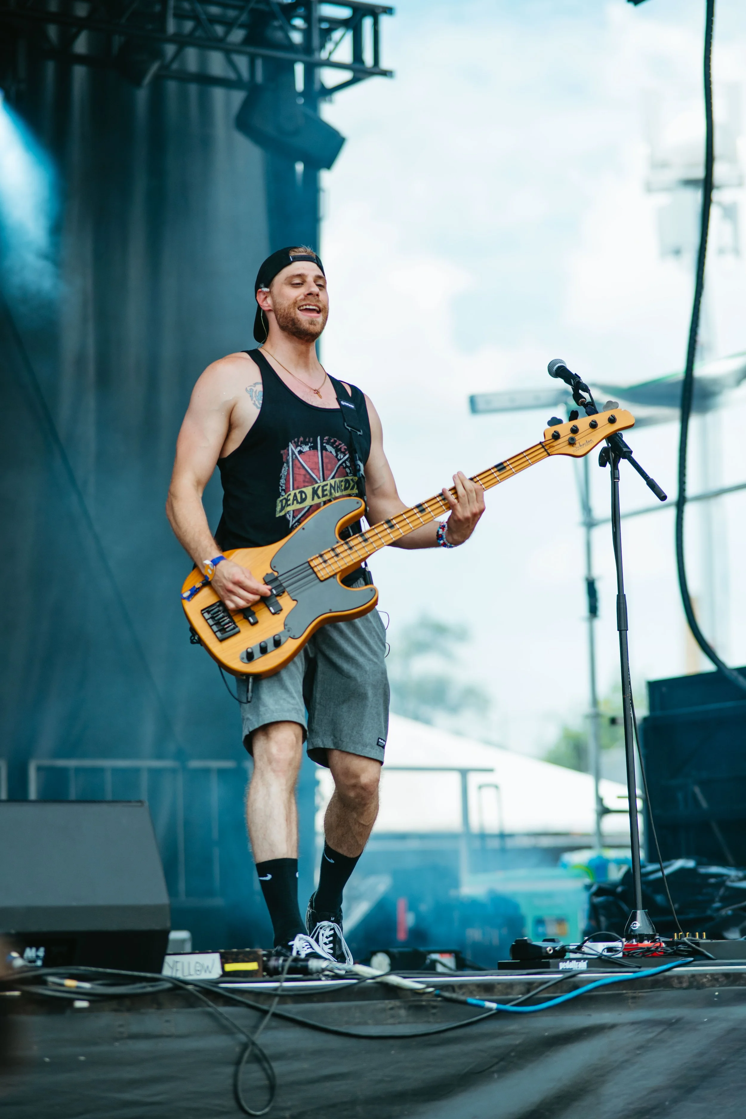 A young man with a beard, wearing a black tank top, shorts, a backwards black cap, and sneakers, is playing an electric bass guitar on stage during a music performance. He is the bass guitarist for the band Flourescents.
