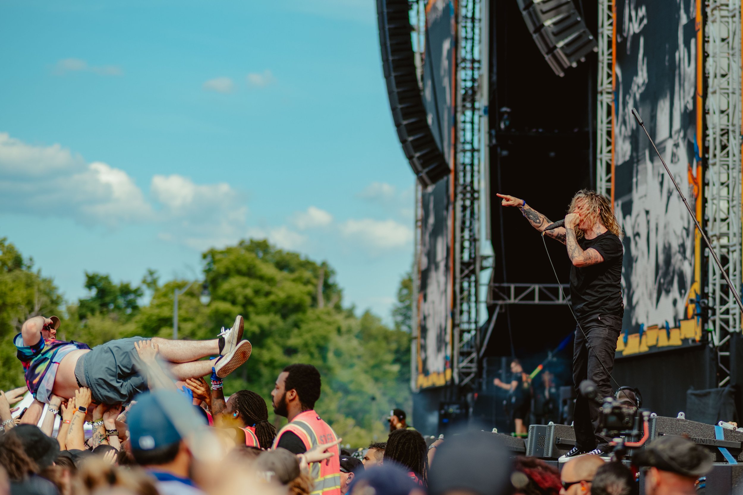 Musician with long hair and tattoos performing on stage at an outdoor concert, with a crowd in front of him, and a stage set against a green background and blue sky.