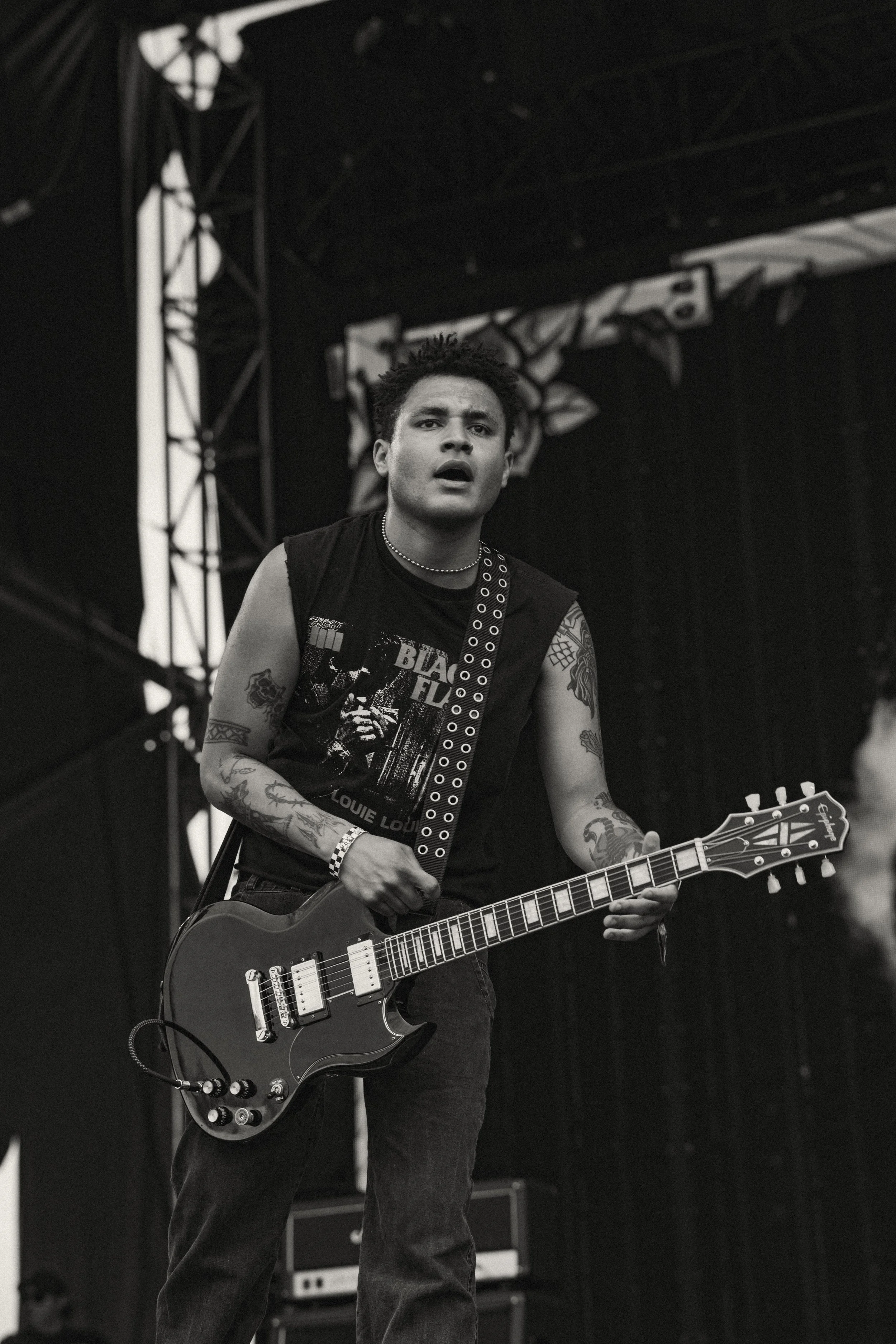 A young man with tattoos on his arms, wearing a sleeveless shirt, playing an electric guitar on stage during a music performance. The scene is in black and white.