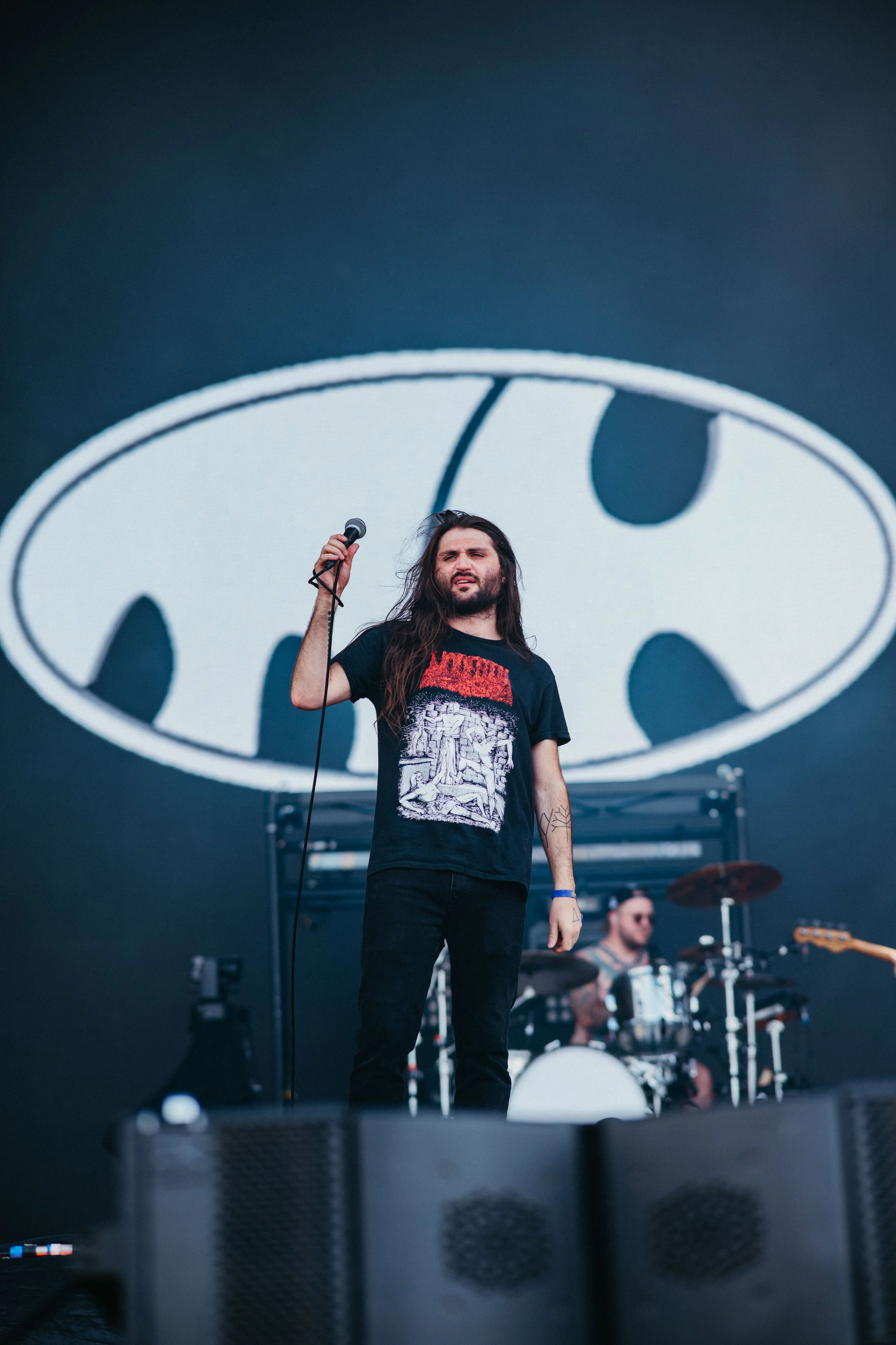 A man with long dark hair and a beard holding a microphone on stage, with a large LED screen behind him displaying the Batman logo. Other band members can be seen playing instruments in the background.