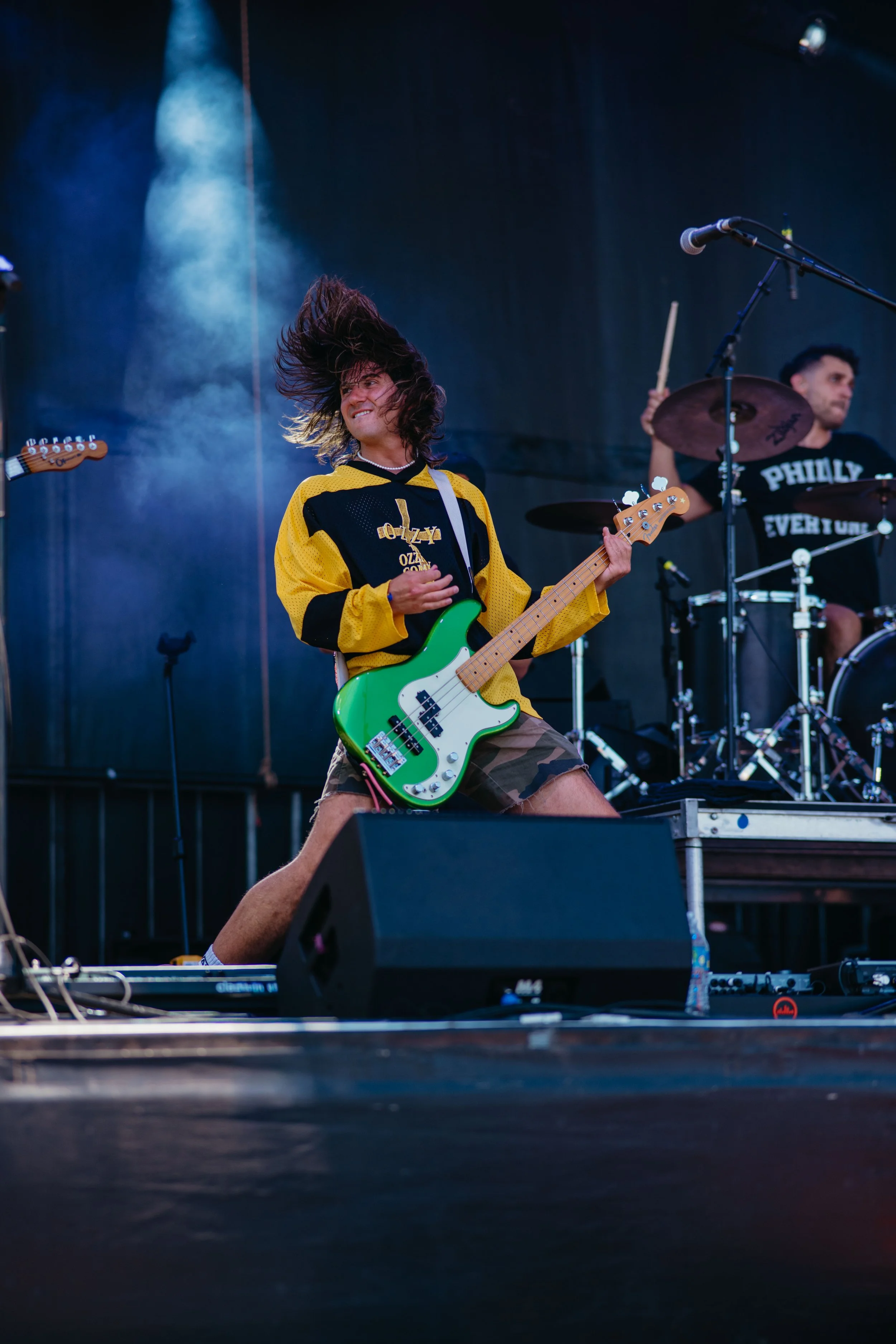 A musician playing a green bass guitar on stage during a concert, with another band member playing drums in the background and stage lighting creating a smoky atmosphere.