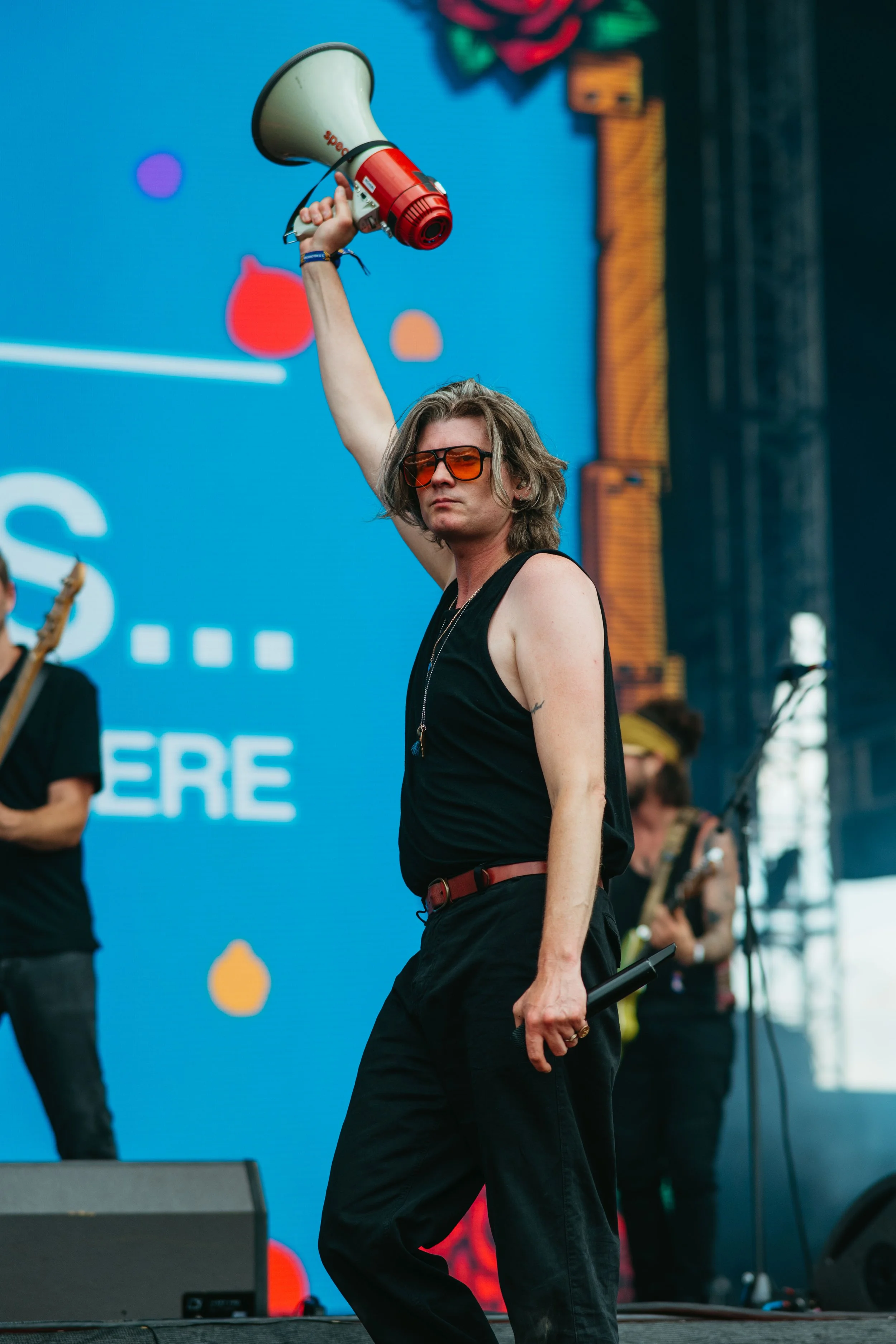 A person with shoulder-length hair and sunglasses holding a megaphone on a stage at a music festival or concert.