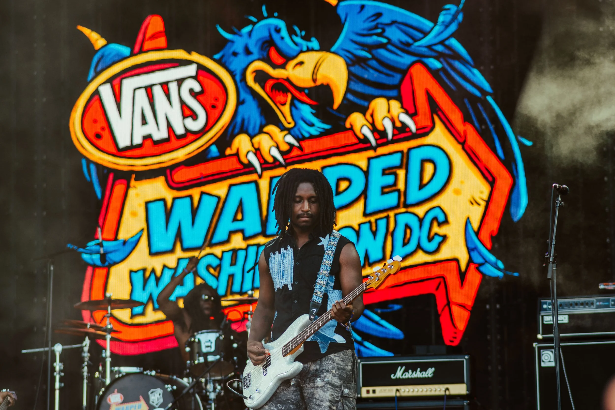 Musician playing guitar on stage at Warped Tour, with large colorful digital screen displaying VANS logo and mascot in the background.