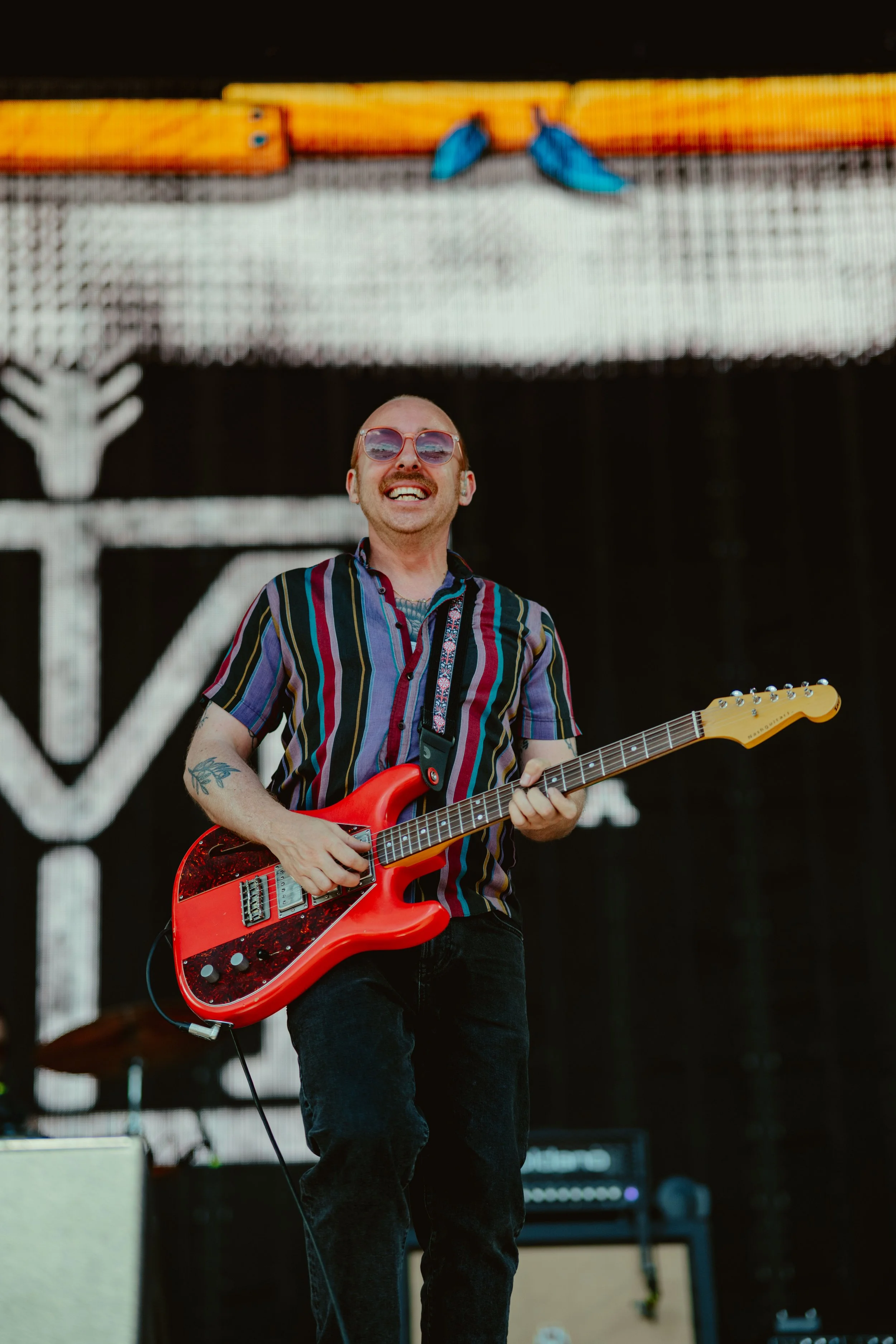 A man with sunglasses and a striped shirt playing an orange electric guitar on stage, with musical equipment in the background.